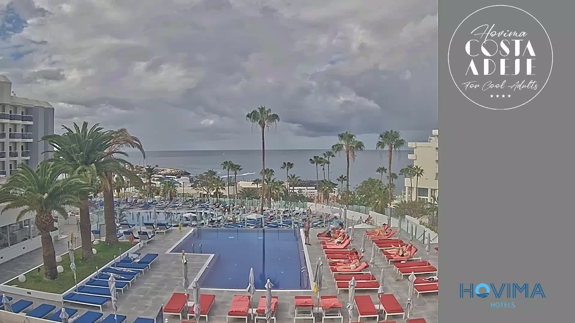 An outdoor pool area with red lounge chairs and palm trees overlooks a calm ocean under a cloudy sky.
