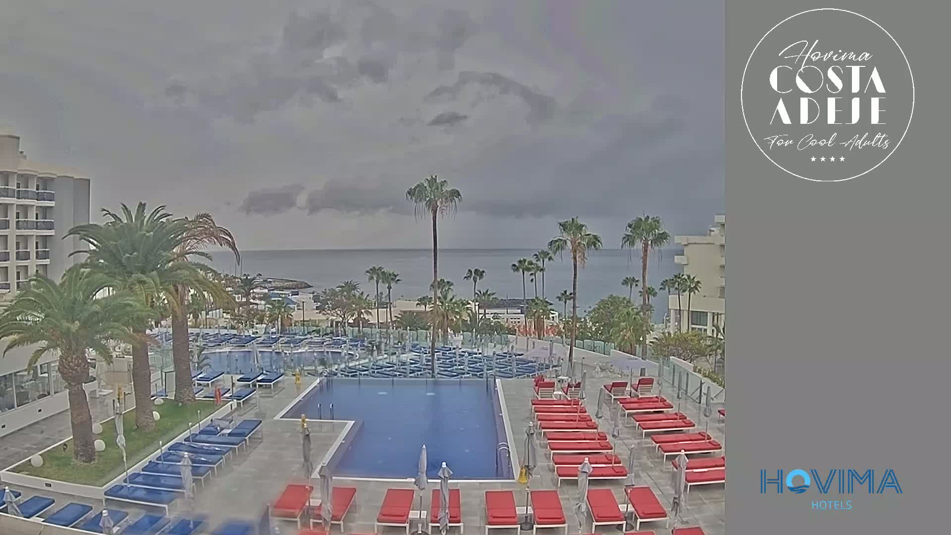 An outdoor pool area with red and blue lounge chairs surrounds a rectangular pool, all situated next to a hotel building with palm trees and an ocean view under a cloudy sky.