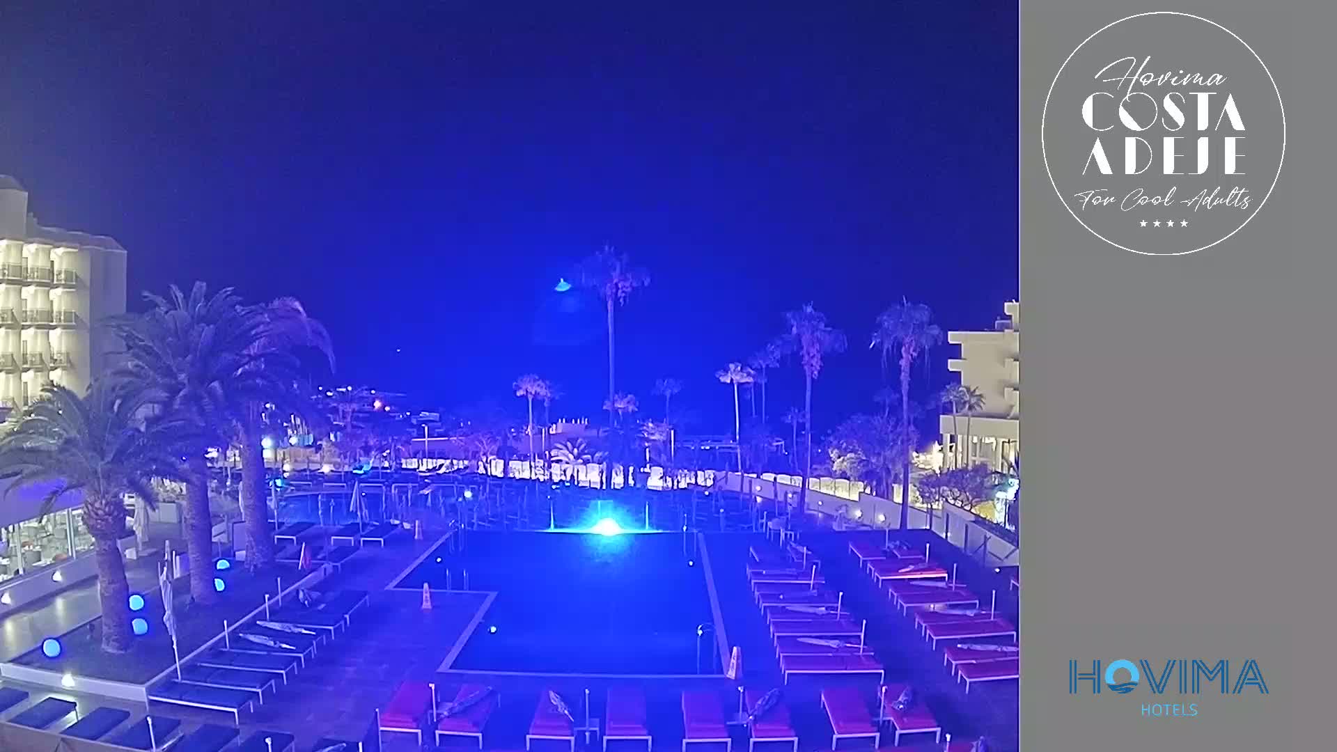 A nighttime view of a resort pool area illuminated in blue light, surrounded by palm trees and hotel buildings under a clear, dark blue sky.