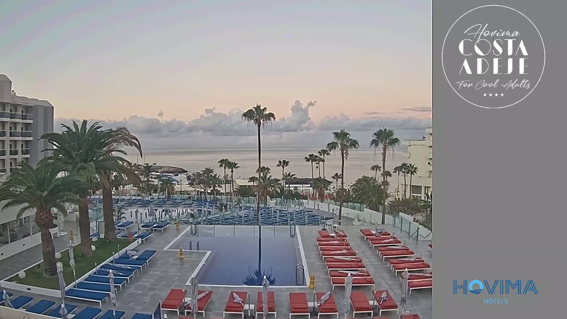 A hotel pool area with red and blue lounge chairs surrounds a swimming pool, overlooking a calm ocean under a partly cloudy sky.