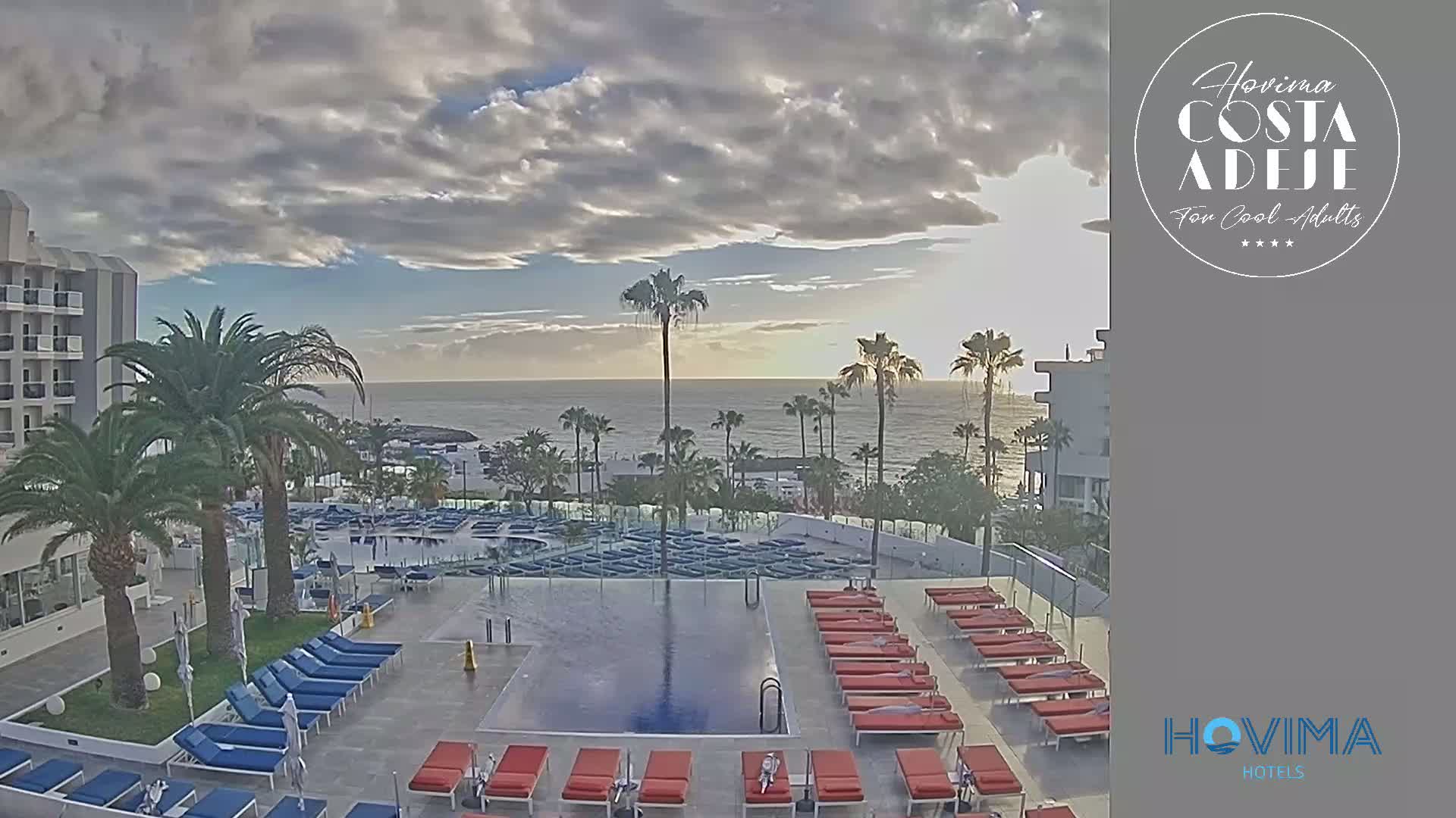 An outdoor pool area with numerous lounge chairs sits adjacent to a hotel overlooking a calm ocean under a partly cloudy sky.