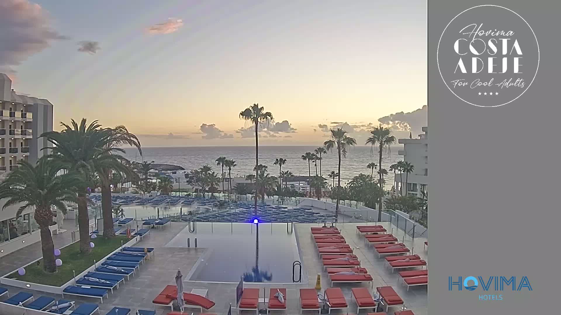 A resort pool area with numerous lounge chairs surrounds a rectangular swimming pool, all overlooking a calm ocean at sunrise.