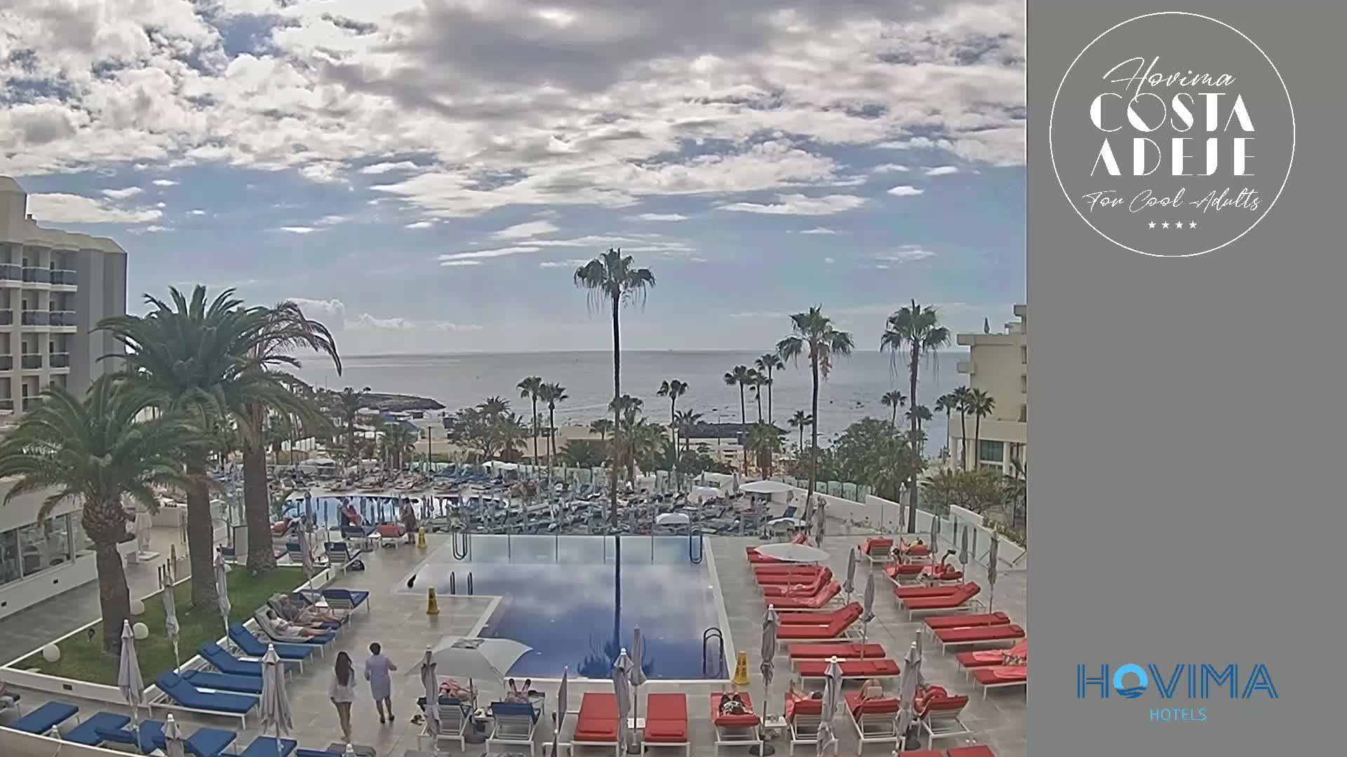 A partly cloudy day reveals a resort pool area with red and blue lounge chairs surrounding a large rectangular pool, palm trees, and a distant ocean view.