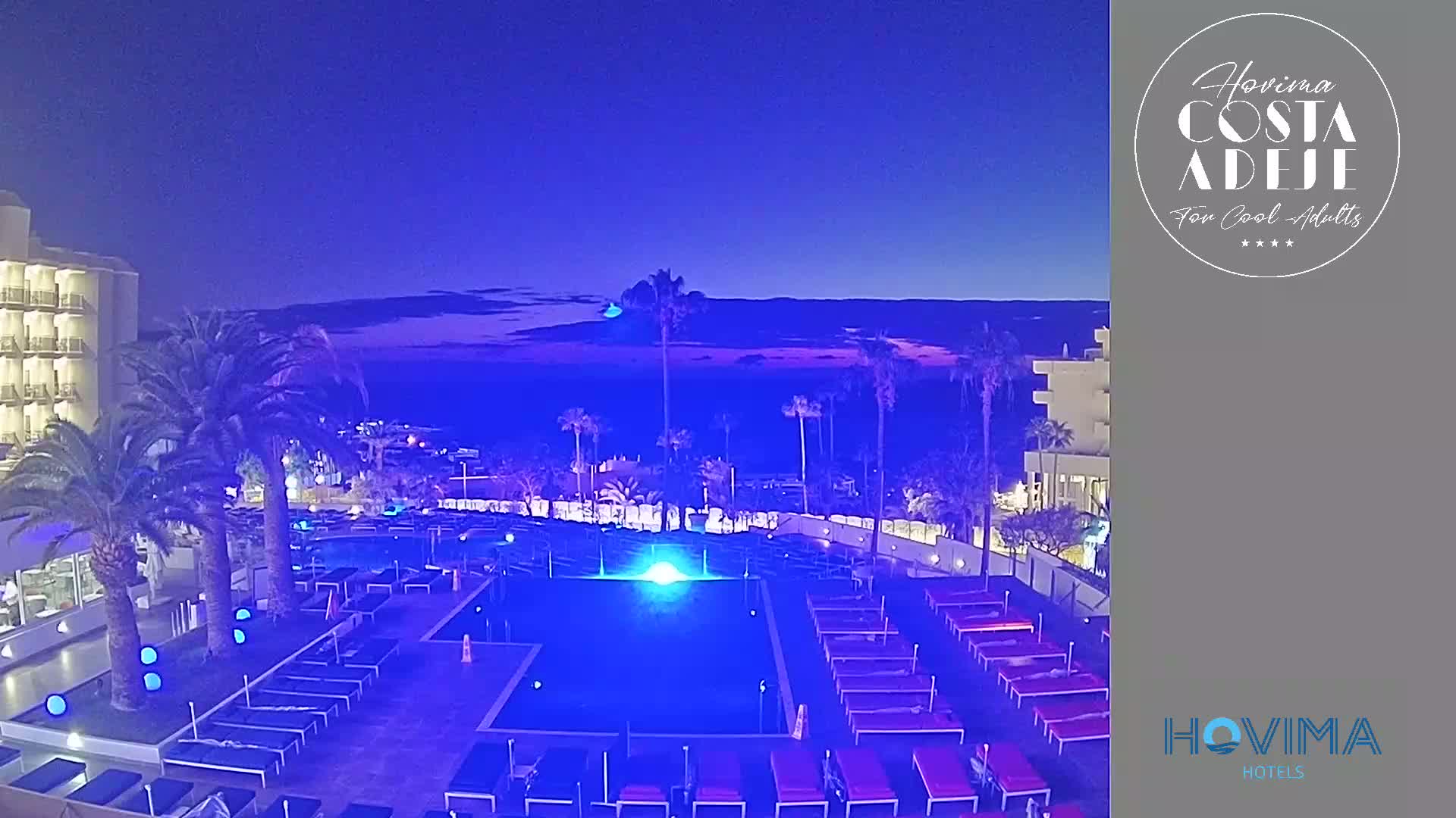 An illuminated resort pool area with lounge chairs and palm trees is situated near a coastline at dusk under a twilight sky.