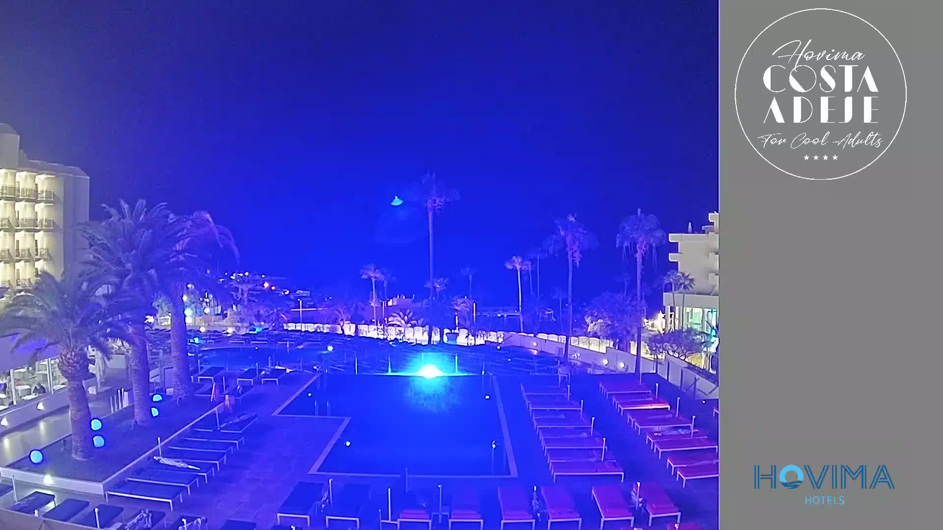 An illuminated swimming pool surrounded by lounge chairs sits at night next to a hotel under a dark blue sky.