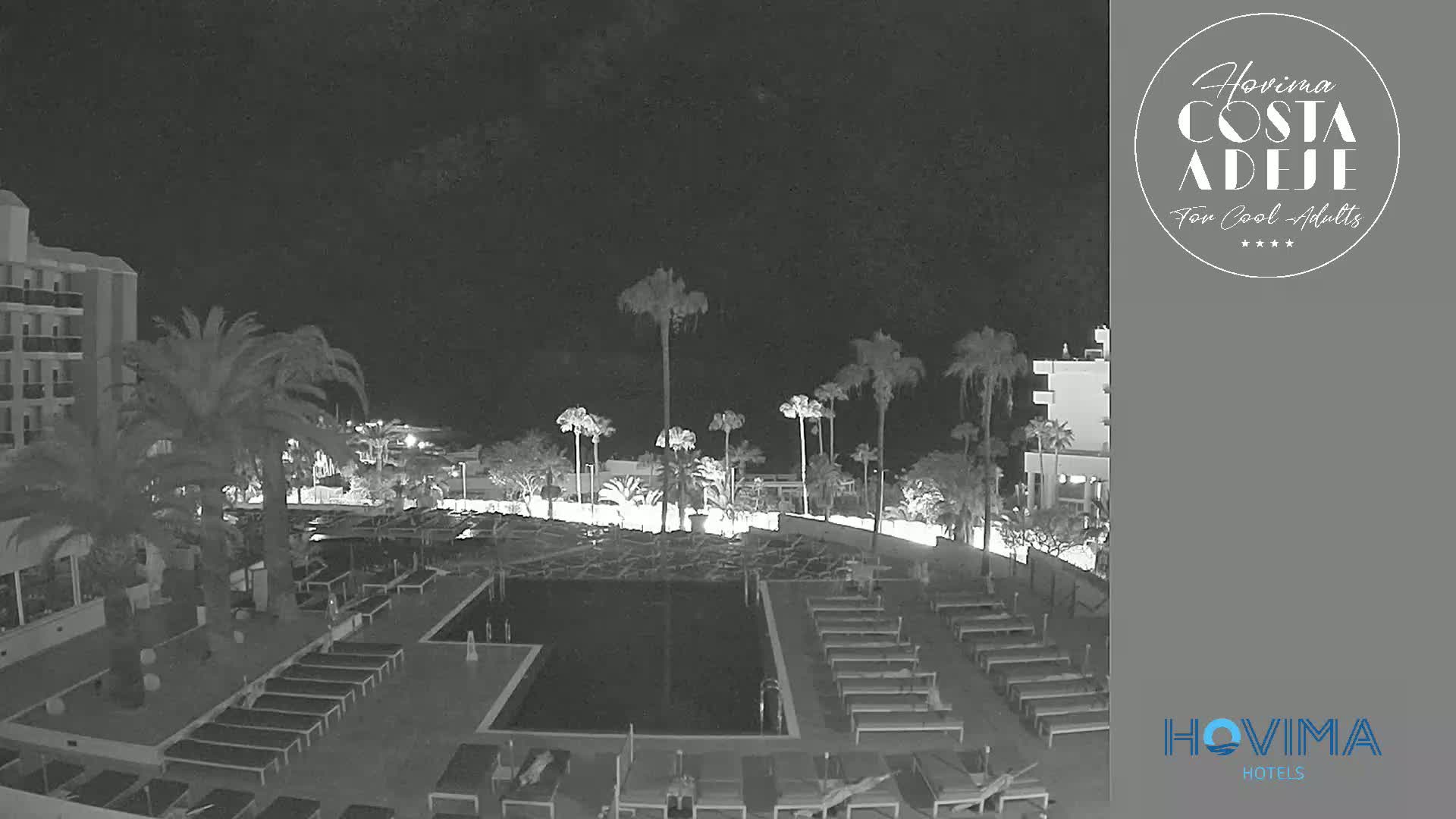 A nighttime view of a hotel pool area with palm trees, lounge chairs, and a building, under a dark sky.