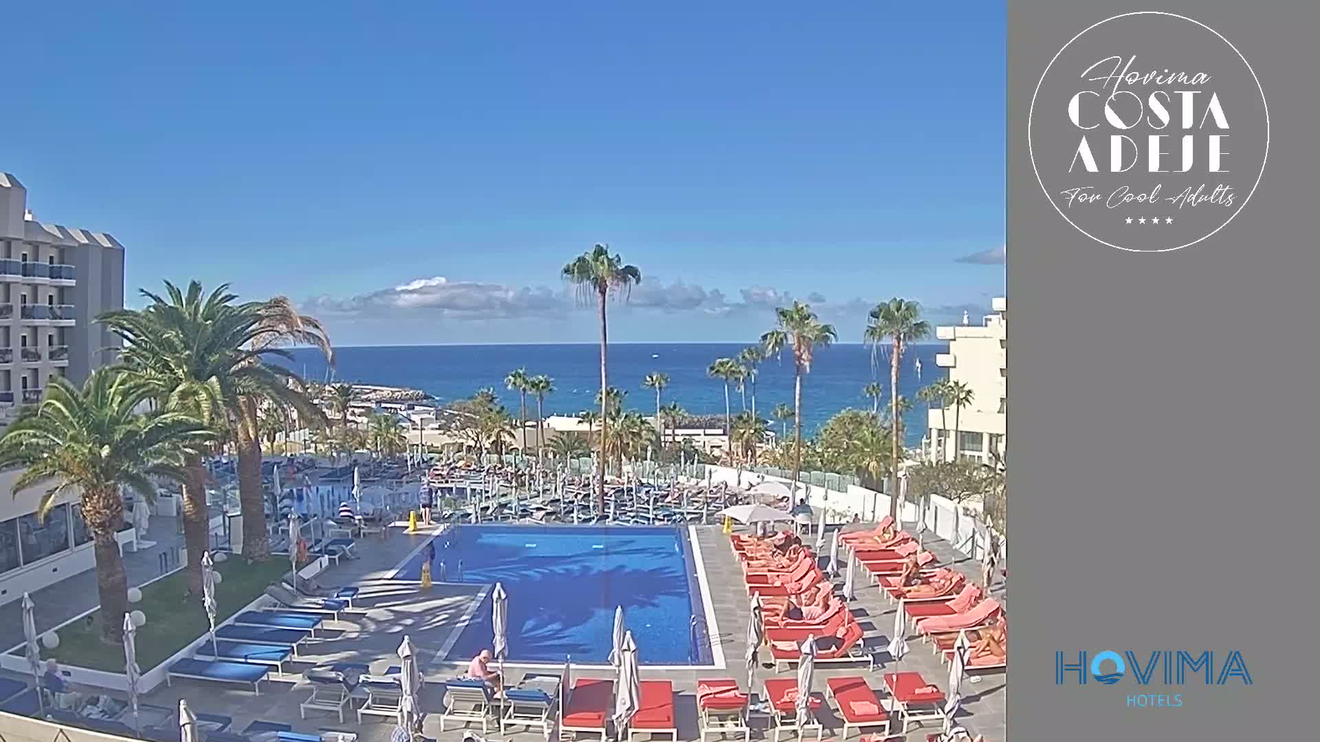 A resort pool area with red lounge chairs surrounding a blue pool, palm trees, and a view of a calm ocean under a mostly sunny sky.