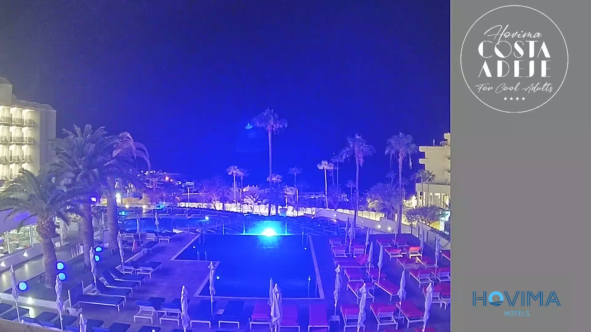A nighttime view of a resort pool area illuminated in blue light, surrounded by palm trees and buildings under a clear, dark sky.