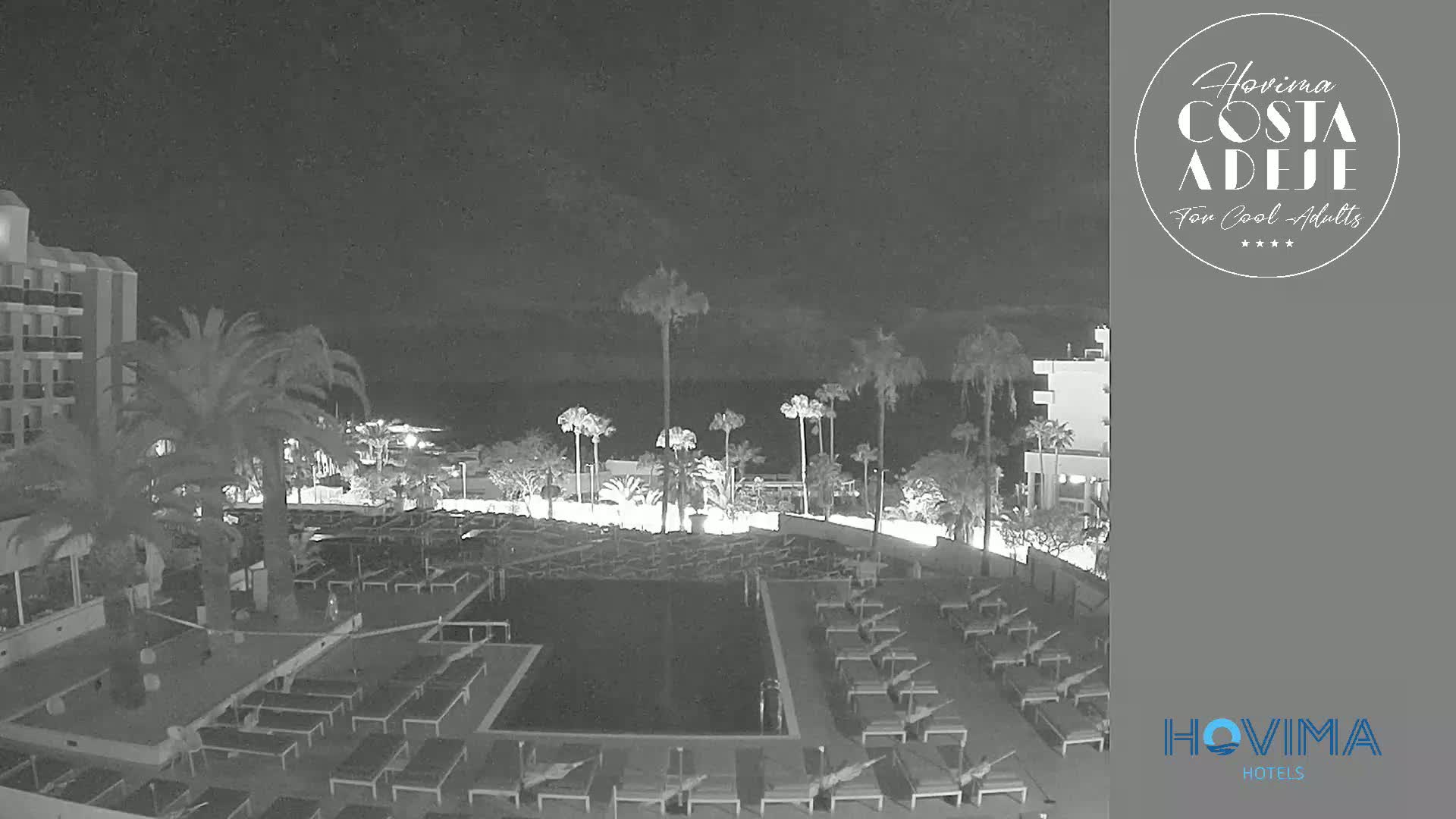A nighttime view of a resort pool area with palm trees, lounge chairs, and a building, under a dark, cloudy sky.
