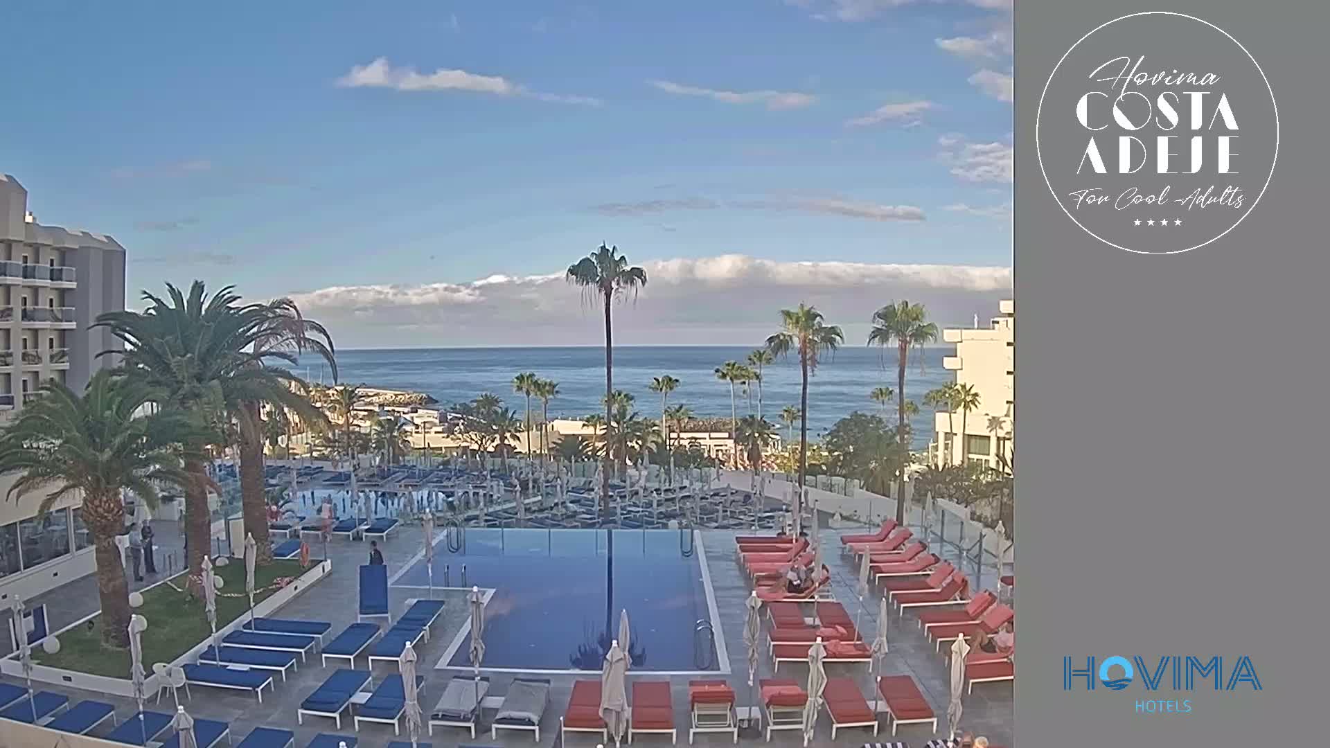 A resort pool with surrounding lounge chairs and palm trees overlooks a calm ocean under a partly cloudy sky.