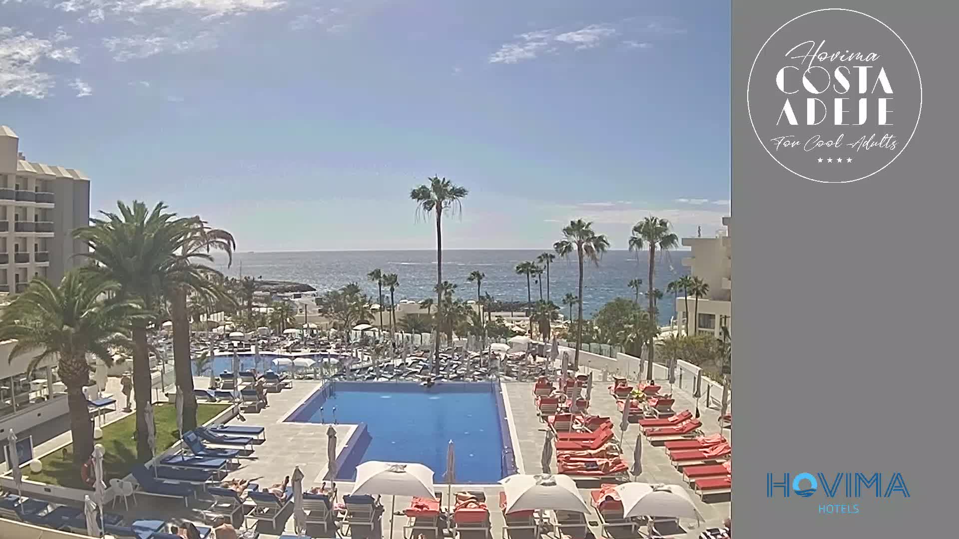 A resort pool area with numerous lounge chairs and umbrellas surrounds a large rectangular pool, all situated beside a coastline under a mostly sunny sky.