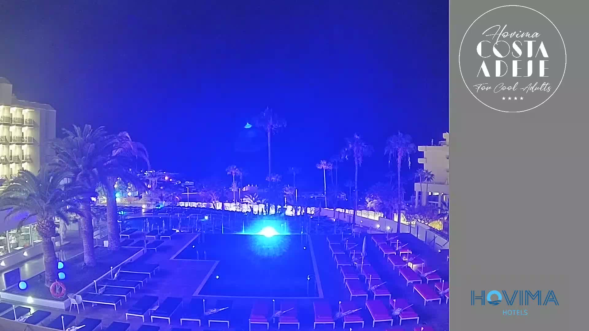 A nighttime view of a hotel pool area, illuminated in blue, surrounded by palm trees and lounge chairs under a dark blue sky.