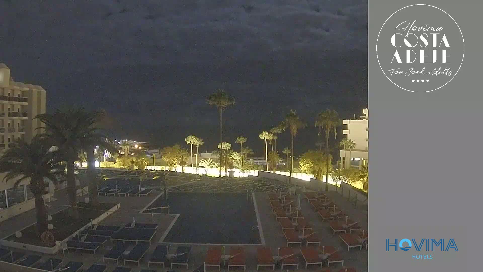 A nighttime view of a resort pool area with palm trees, lounge chairs, and a multi-story hotel building under a dark, cloudy sky.