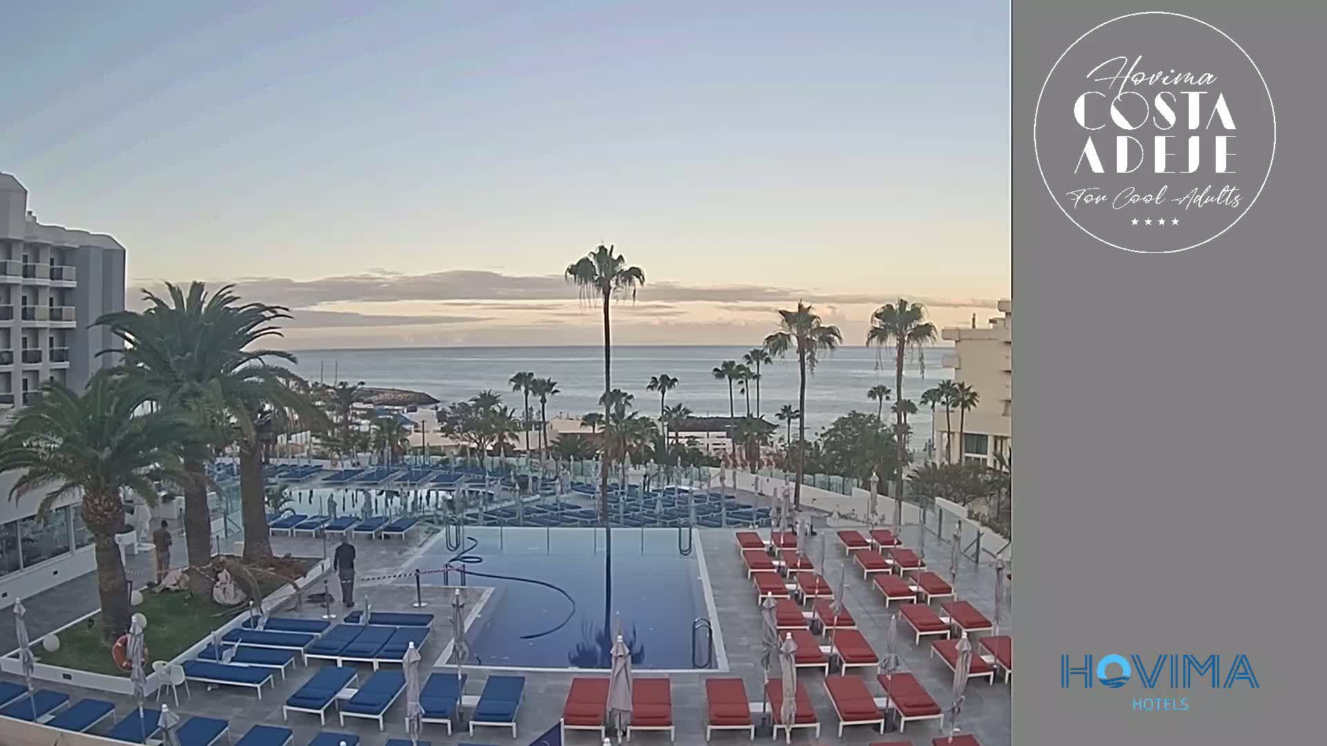 A resort pool area with many lounge chairs surrounds a rectangular pool, overlooking a calm ocean under a clear, dawn sky.