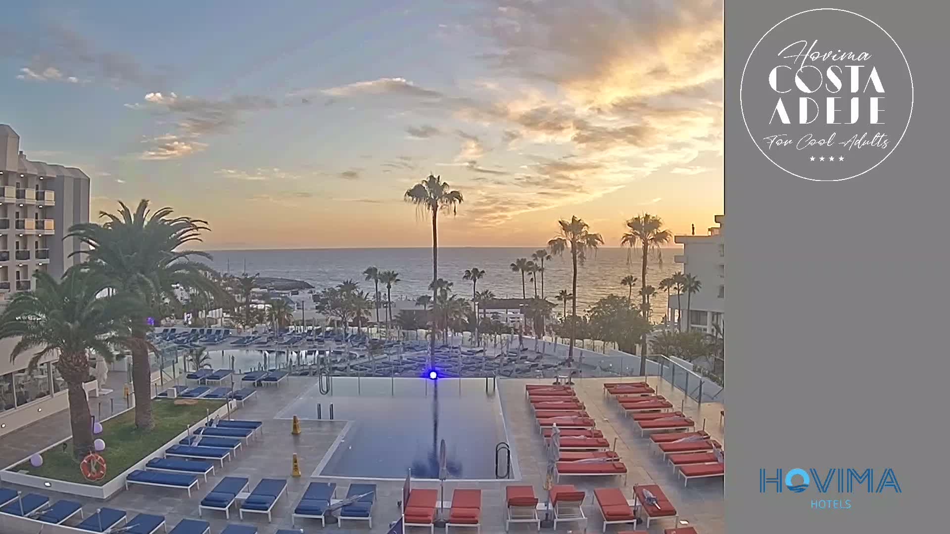 An outdoor pool area with numerous lounge chairs surrounds a rectangular pool, overlooking a calm ocean at sunset, with palm trees lining the perimeter.