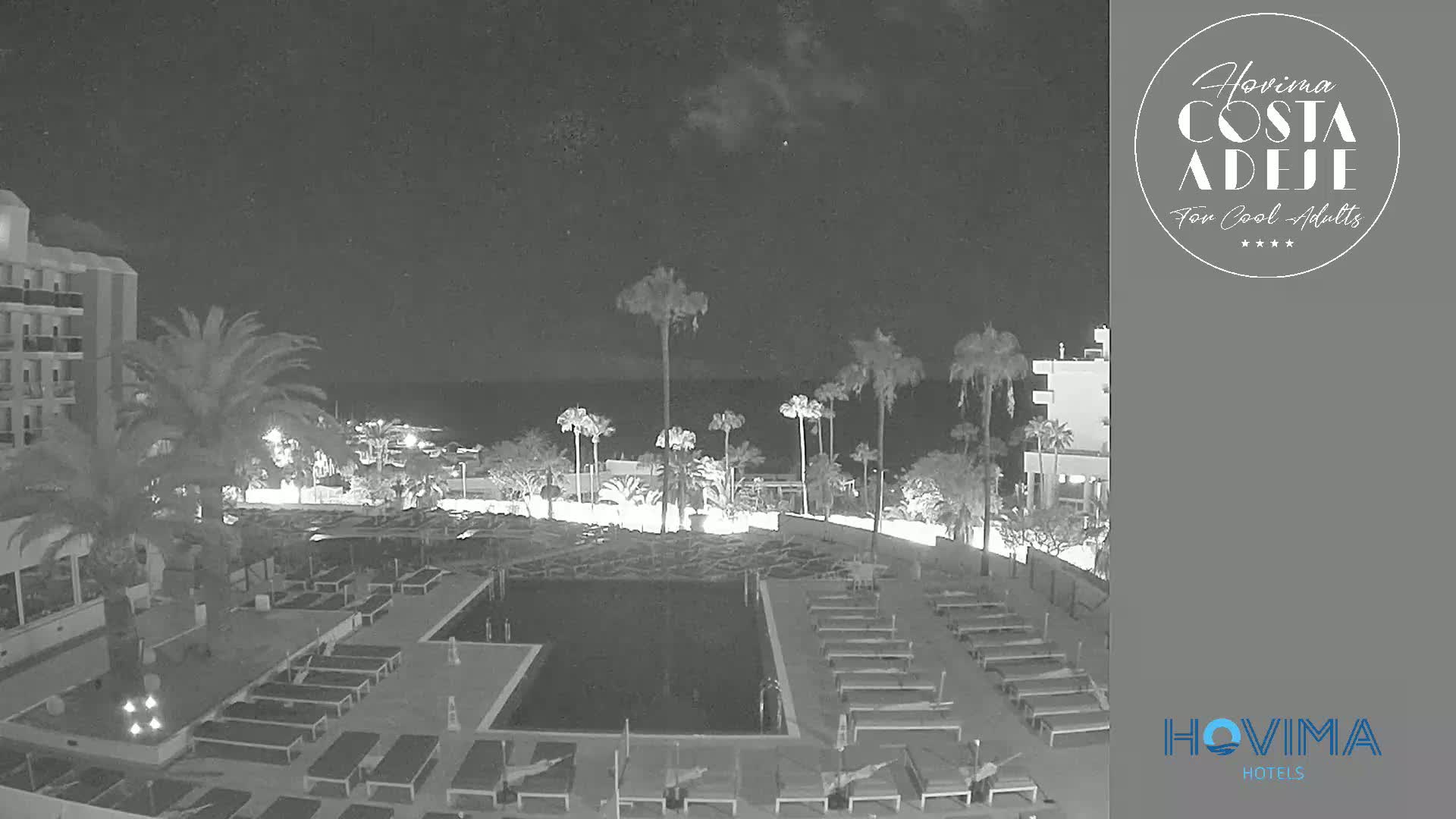 A nighttime, low-light view of a resort pool area with palm trees, lounge chairs, and a building, overlooking a dark ocean.