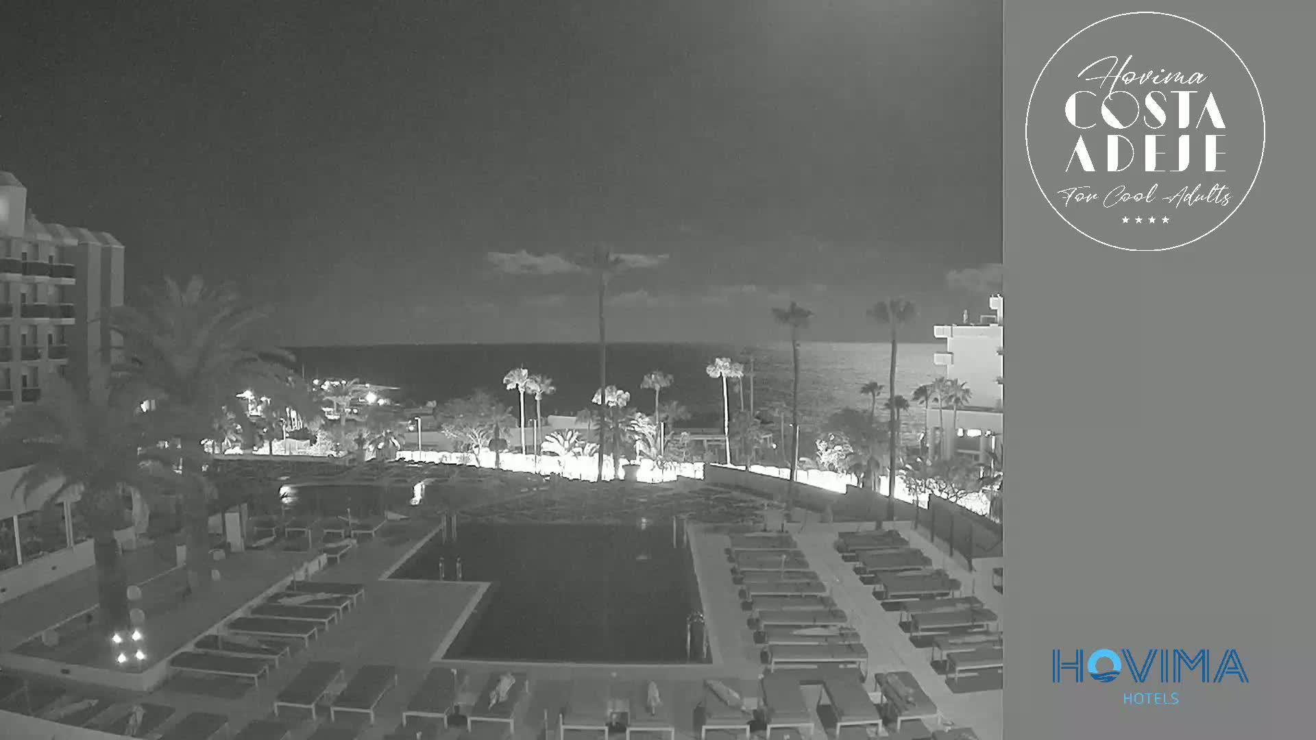 A nighttime view of a resort pool area with palm trees and a calm ocean in the background.