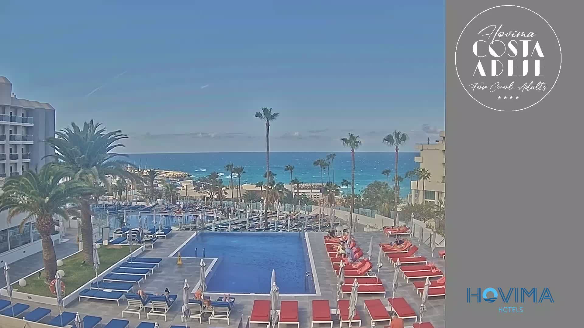 A hotel pool area with red lounge chairs and umbrellas overlooks a calm ocean under a clear blue sky.