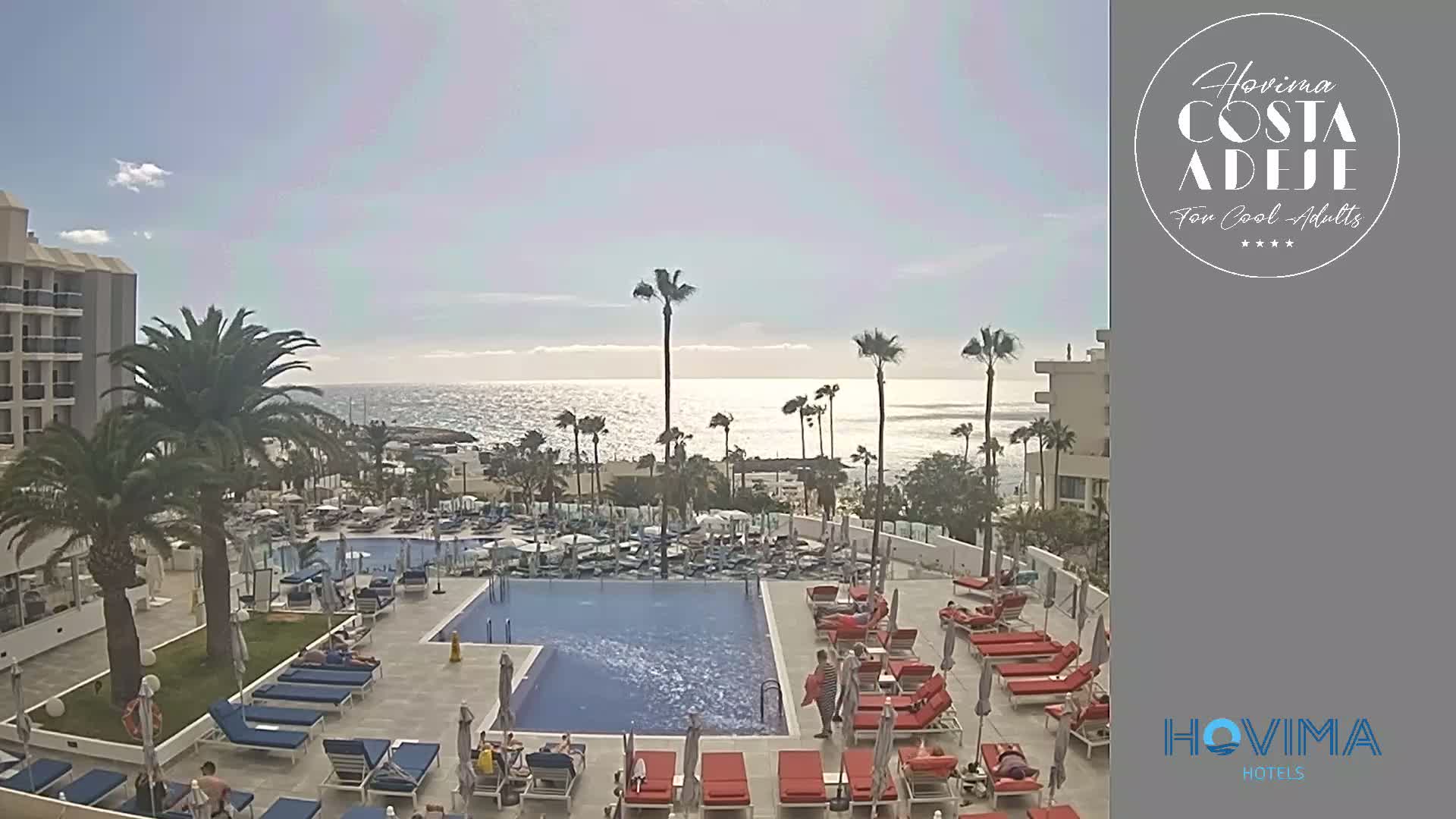 A resort pool area with red lounge chairs surrounding a large rectangular pool, palm trees, and a building overlooking a calm ocean under a mostly sunny sky.
