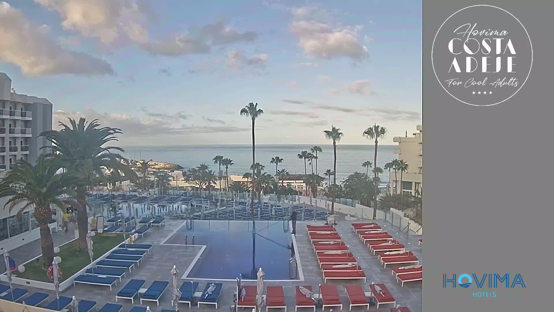 A hotel pool area with numerous empty lounge chairs surrounds a rectangular pool, overlooking a calm ocean under a partly cloudy sky.