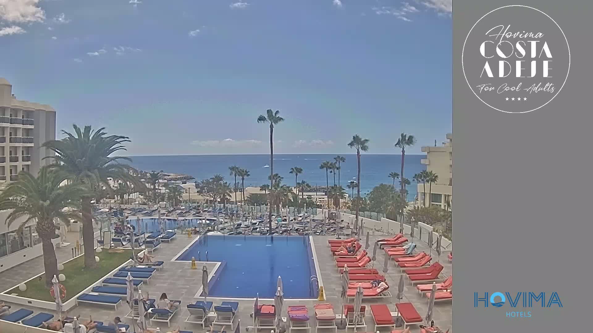 A resort pool area with lounge chairs and palm trees overlooks a calm ocean under a mostly sunny sky.