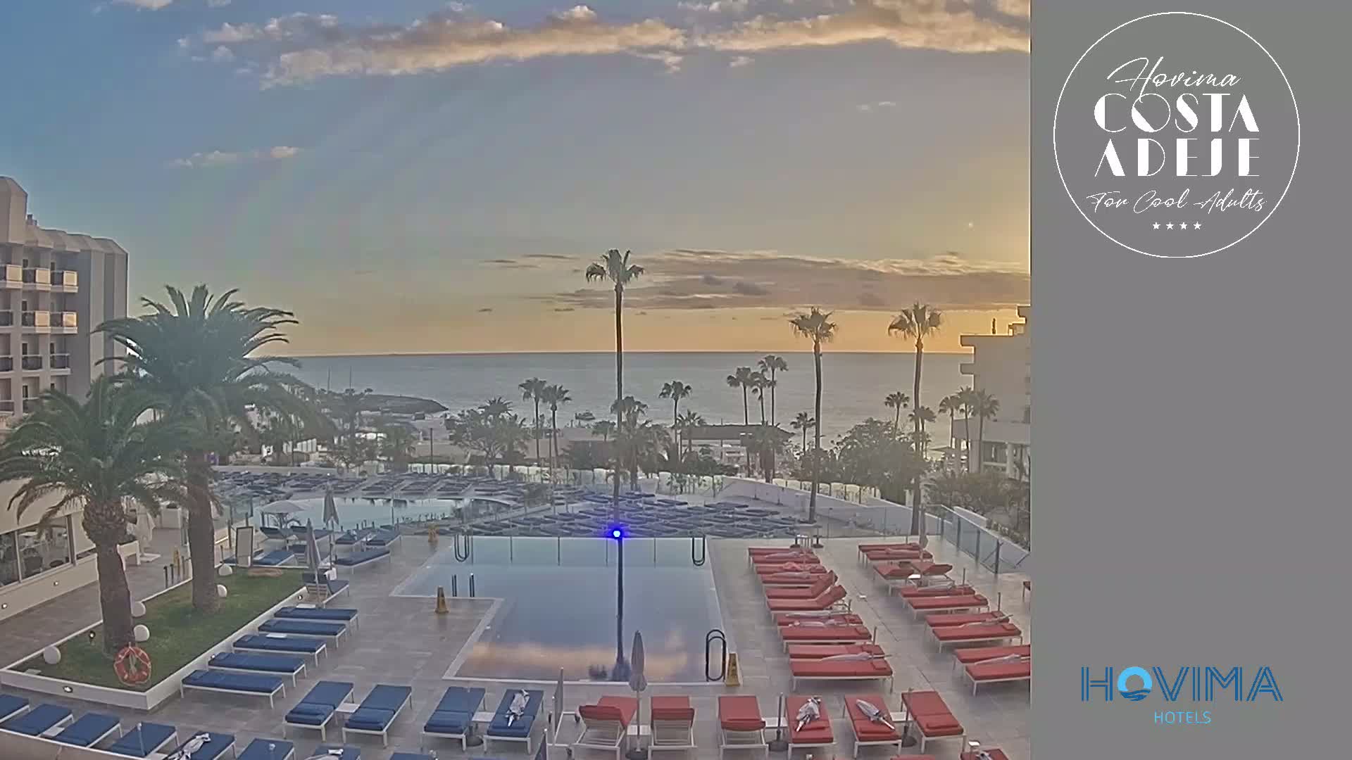 An outdoor pool area with numerous lounge chairs surrounding it overlooks a calm ocean under a partly cloudy sunset sky.