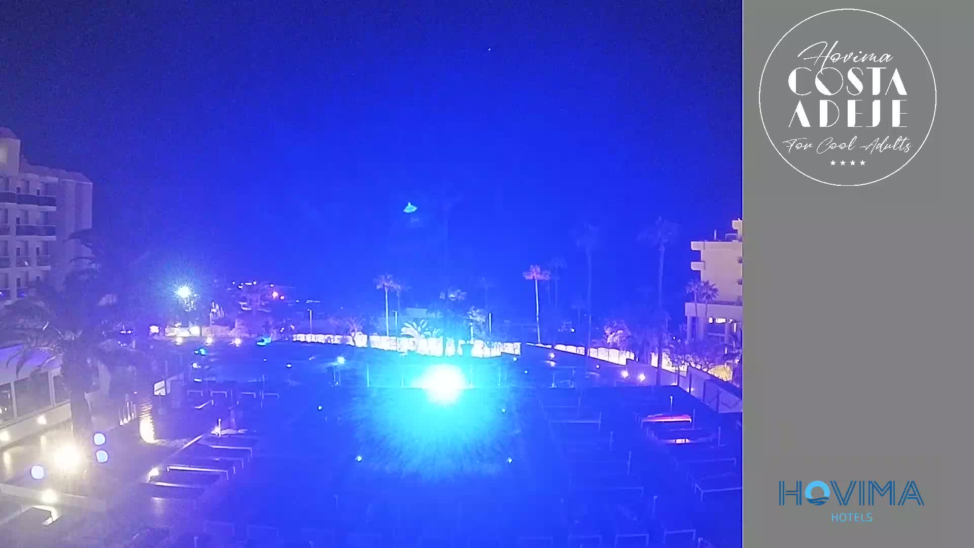 A nighttime view of a resort pool area, illuminated by blue lights, under a clear, dark blue sky.