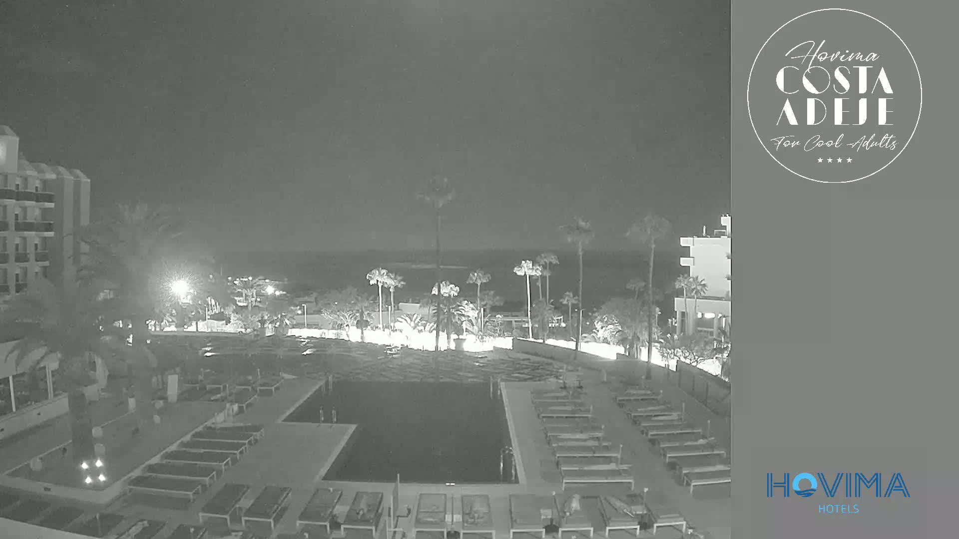 A nighttime, low-light view of a resort pool area with palm trees, lounge chairs, and buildings surrounding it.