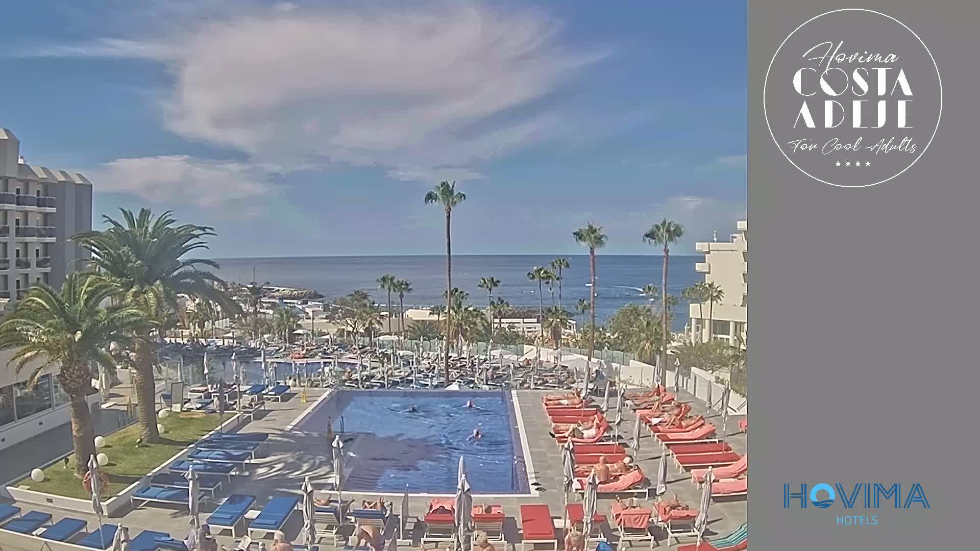 A resort pool area with sunbathers and swimmers overlooks a calm ocean under a partly cloudy sky.