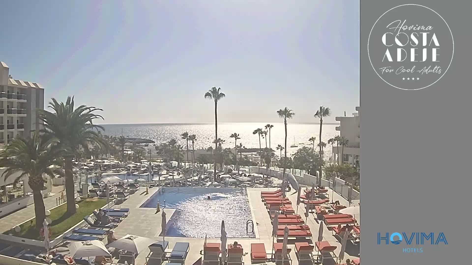 A resort pool area with many lounge chairs and umbrellas surrounds a large swimming pool, overlooking a calm ocean on a sunny day.