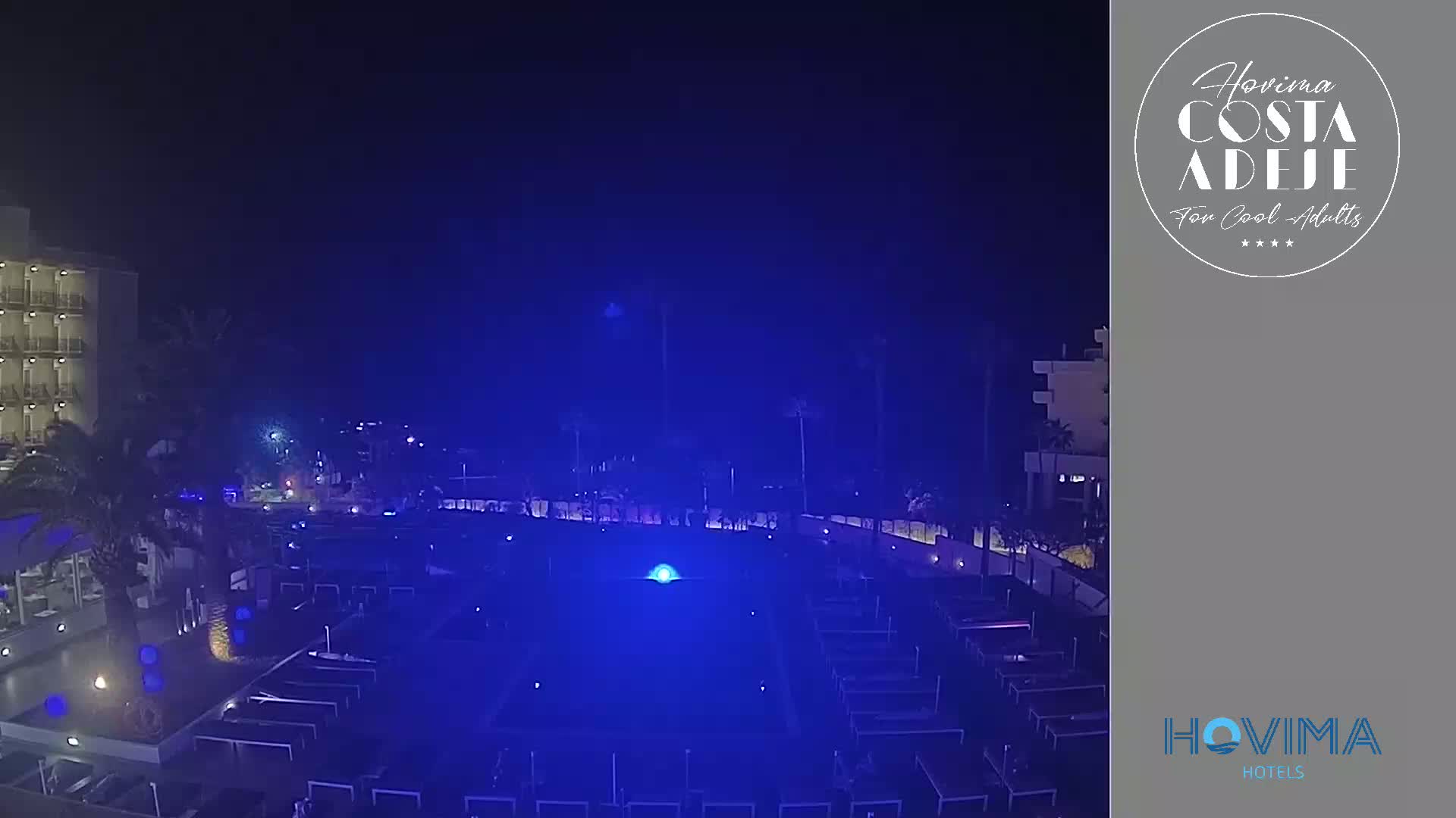 A nighttime view of a resort pool area illuminated in blue, with a building in the background under a dark, clear sky.