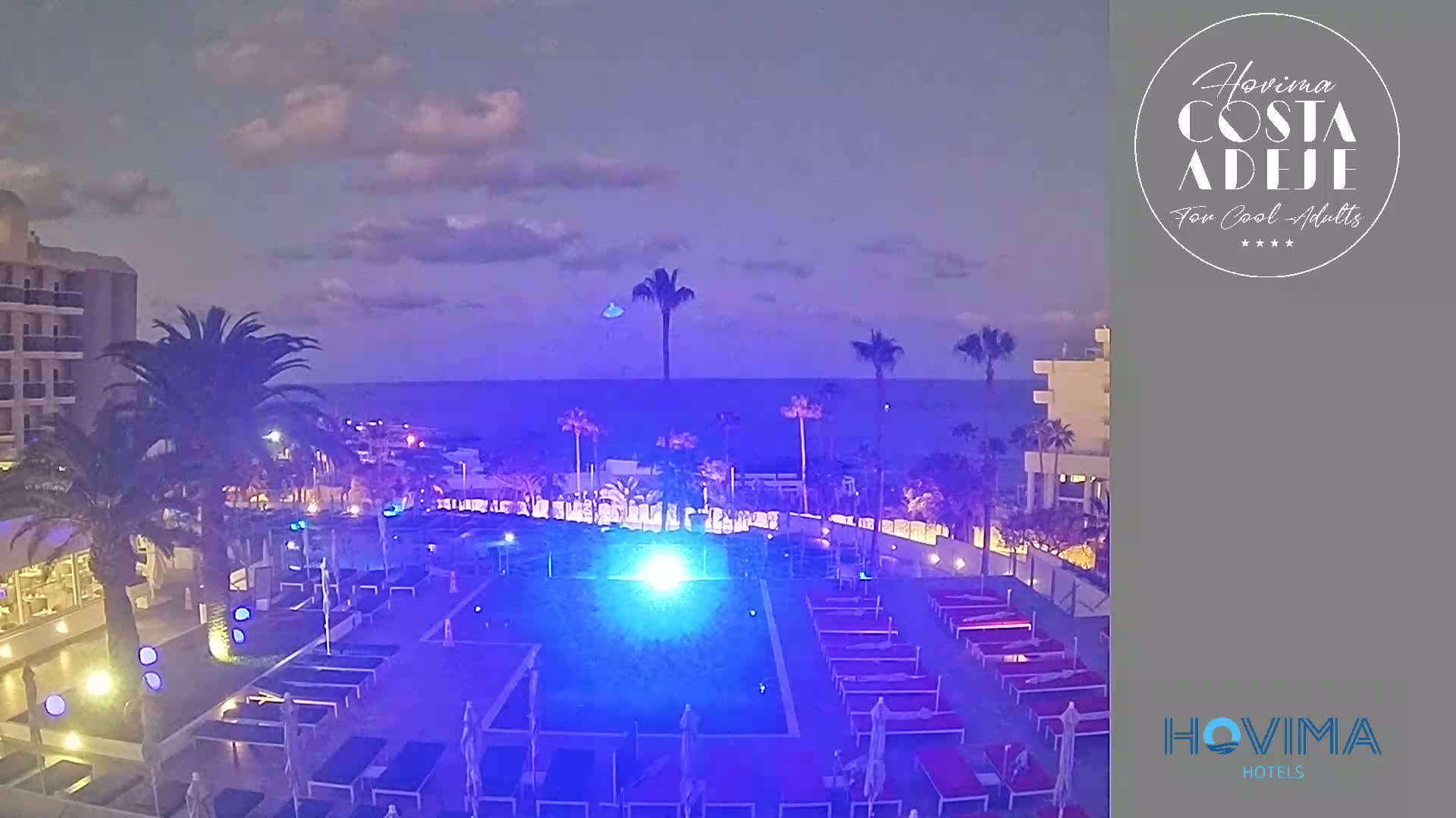 A nighttime view of a resort pool area illuminated in blue light, overlooking a calm ocean under a partly cloudy sky.