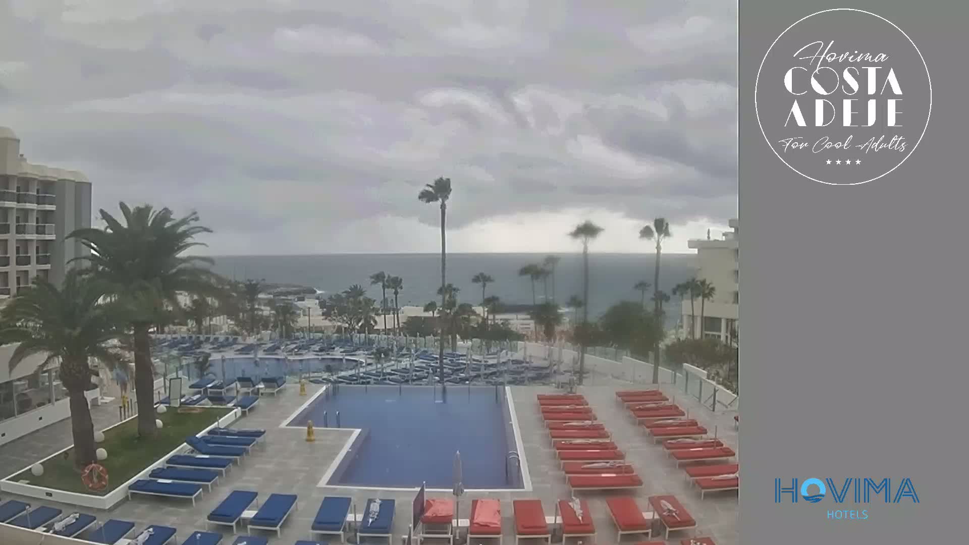 An outdoor swimming pool area with lounge chairs and a large hotel building is visible, under an overcast, windy sky, overlooking a calm ocean.