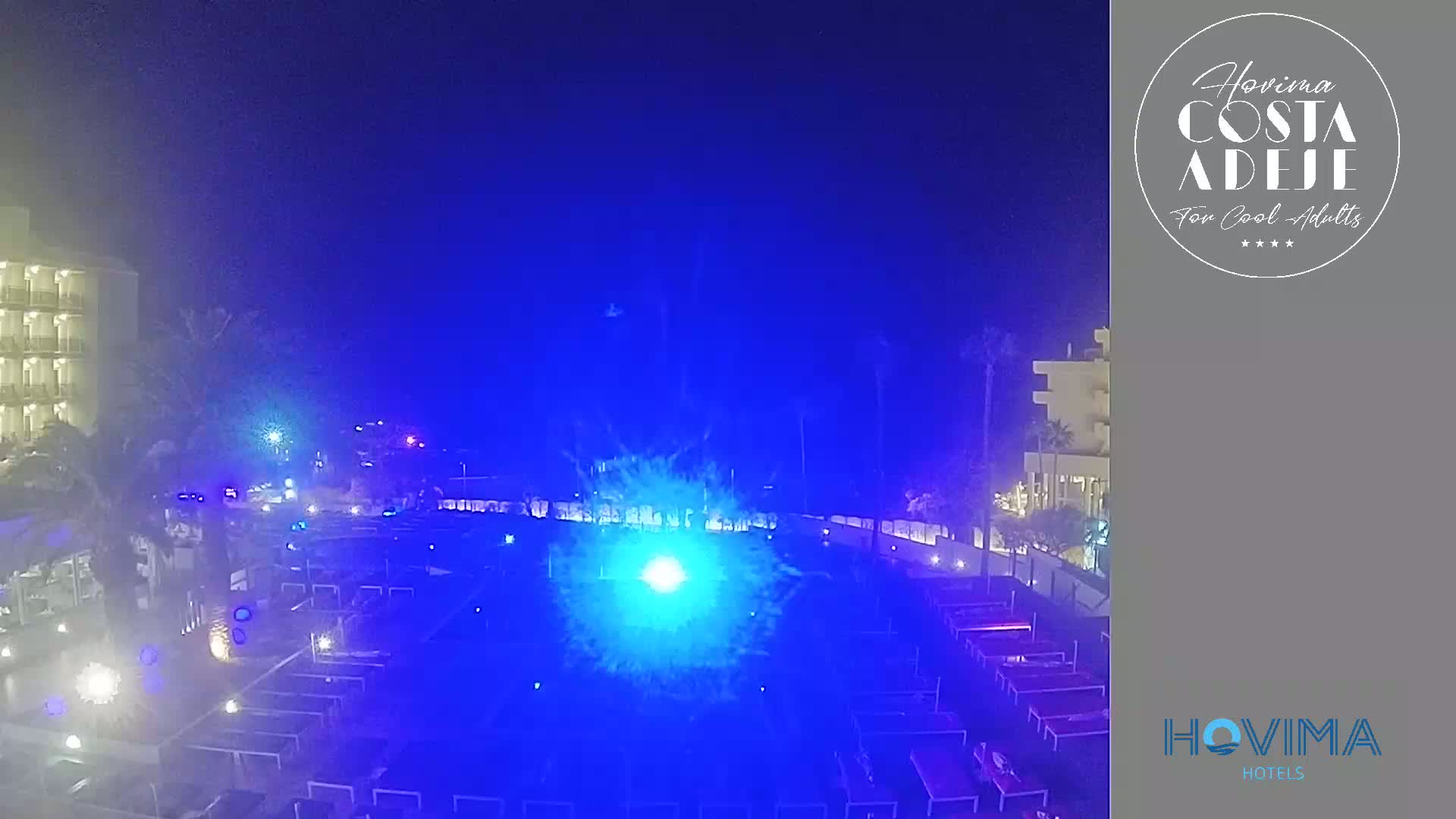 A nighttime view of a resort area with a bright blue light show illuminating an outdoor space filled with lounge chairs and tables.