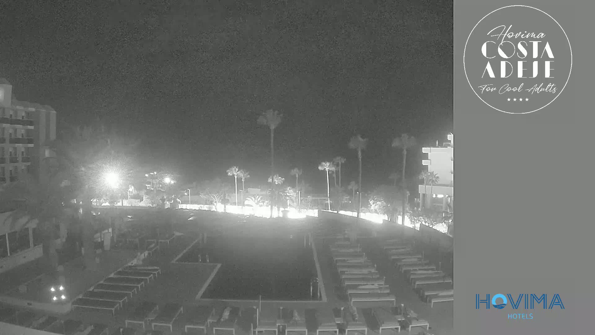 A nighttime, black-and-white view of a resort pool area with palm trees and hotel buildings under a clear, dark sky.