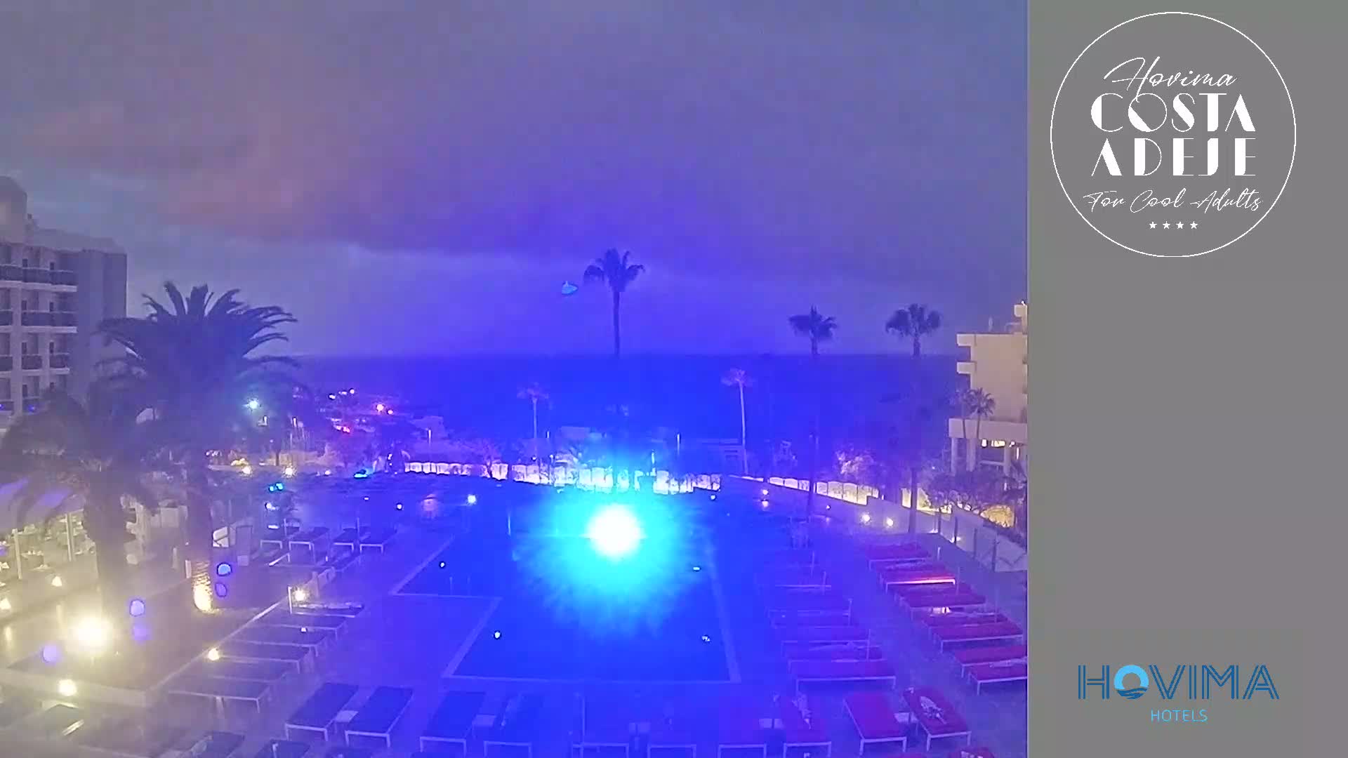 An illuminated swimming pool at a resort next to the ocean under a stormy, twilight sky.