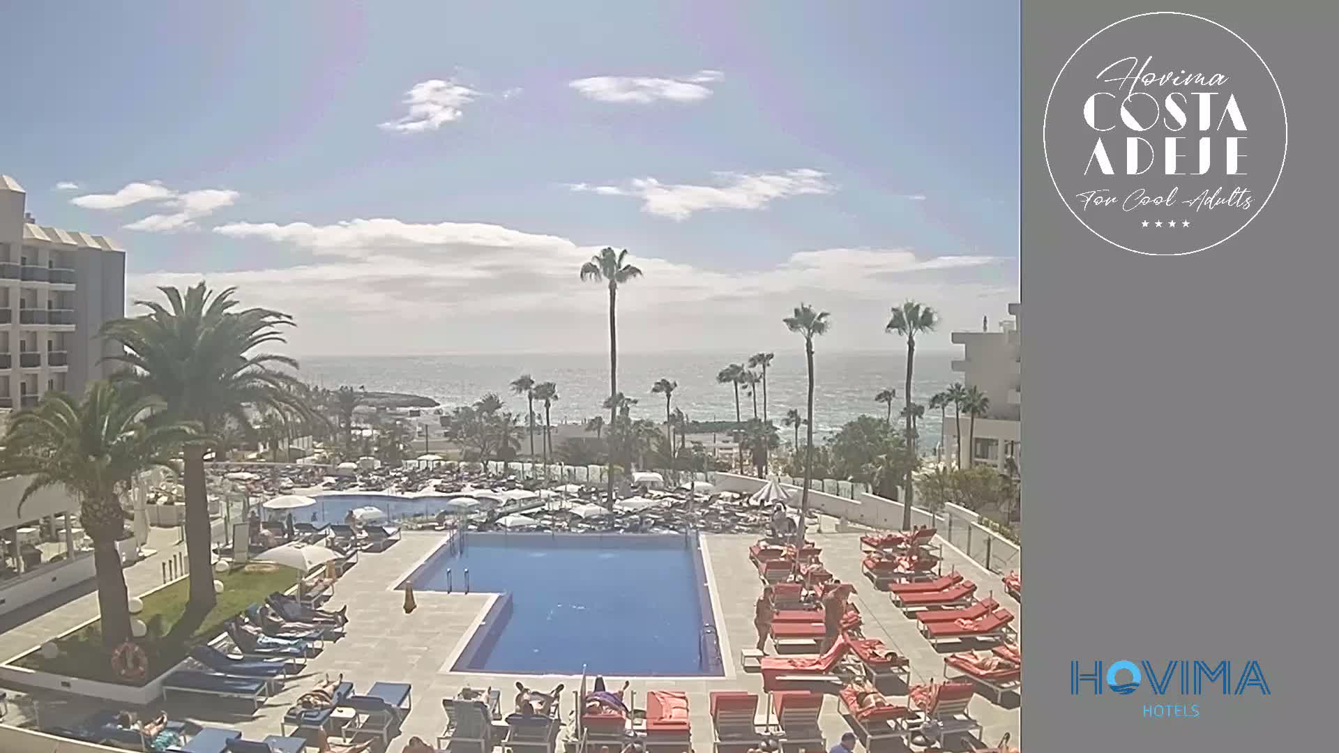 A resort pool area with numerous lounge chairs and sunbathers is situated next to a large hotel, overlooking a calm ocean under a mostly sunny sky.