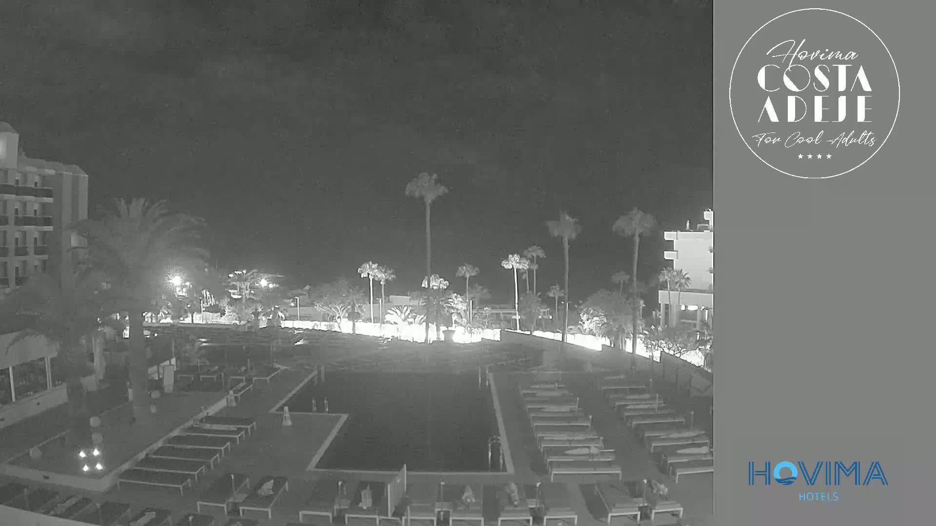 A nighttime view of a resort pool area with palm trees, lounge chairs, and buildings under a dark, clear sky.