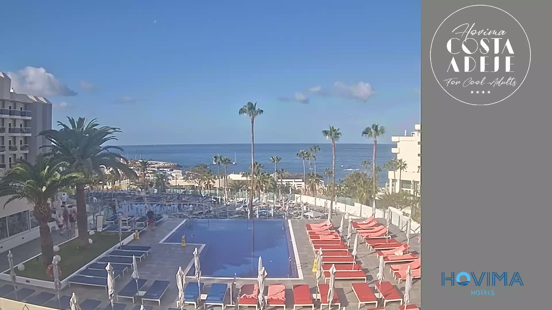 A resort pool area with lounge chairs and palm trees, overlooking a calm ocean under a mostly sunny sky.