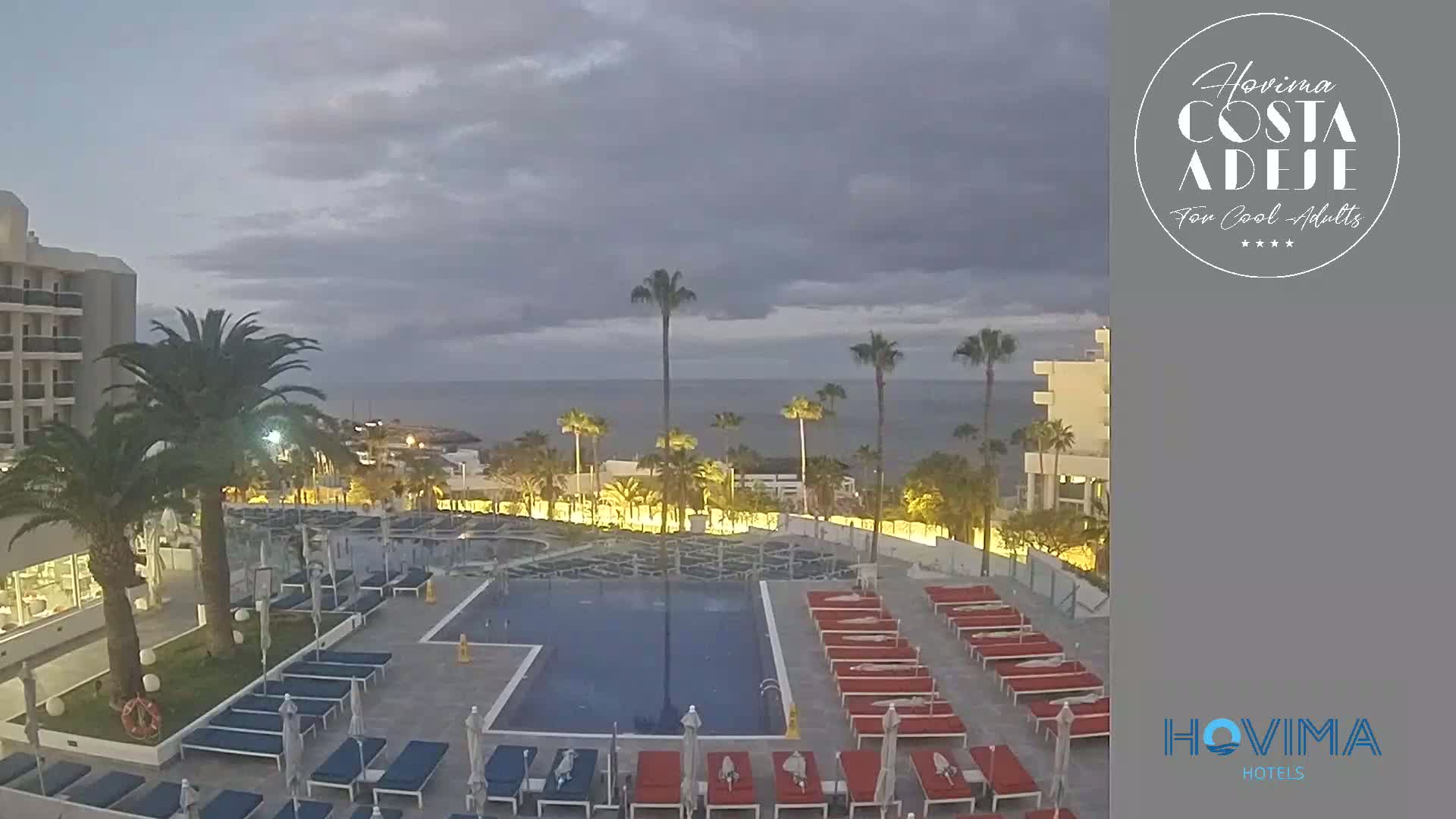 An outdoor pool area with lounge chairs and palm trees, situated near a calm ocean under a cloudy twilight sky.