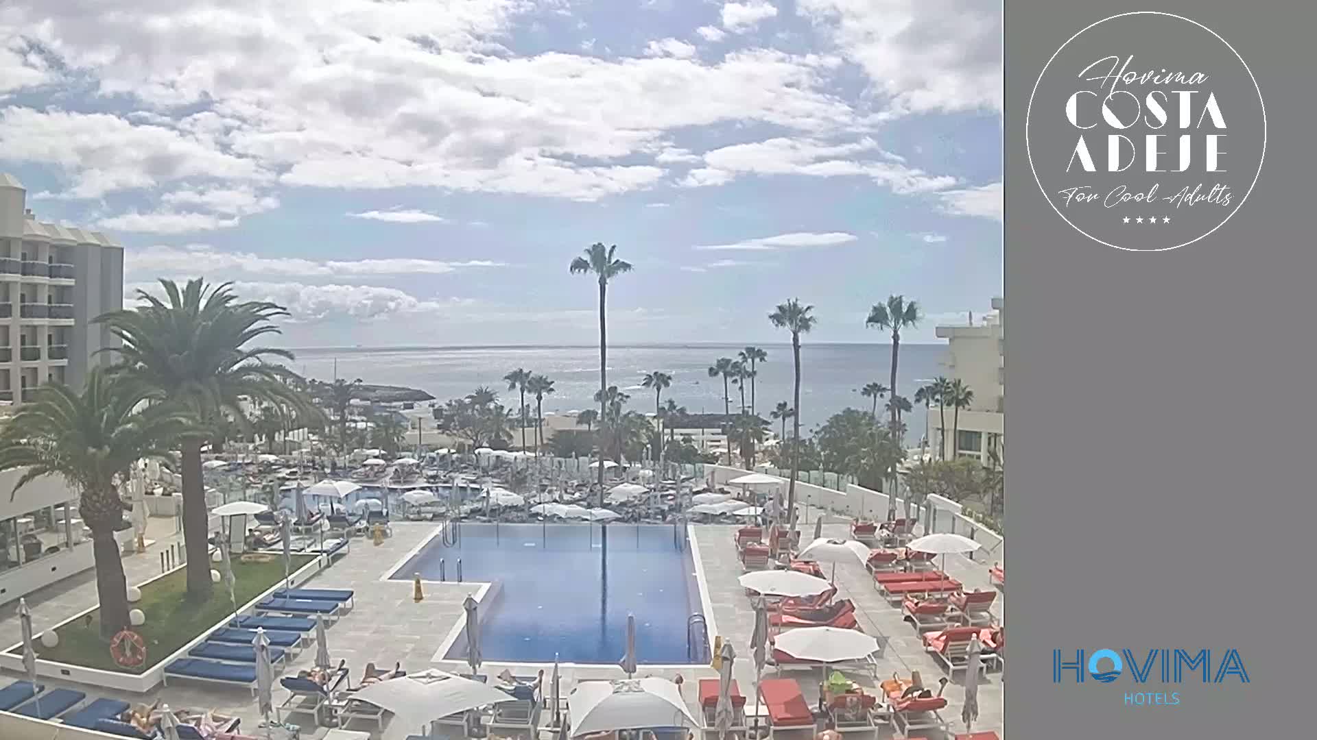 A resort pool area with lounge chairs and umbrellas overlooks a calm ocean under a partly cloudy sky.