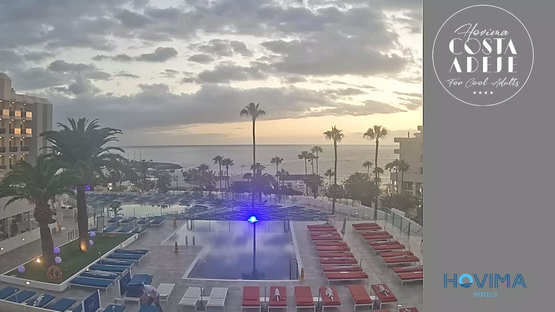 An outdoor pool area with numerous lounge chairs surrounding a pool and a hotel building in the background, under a partly cloudy sky at sunset.