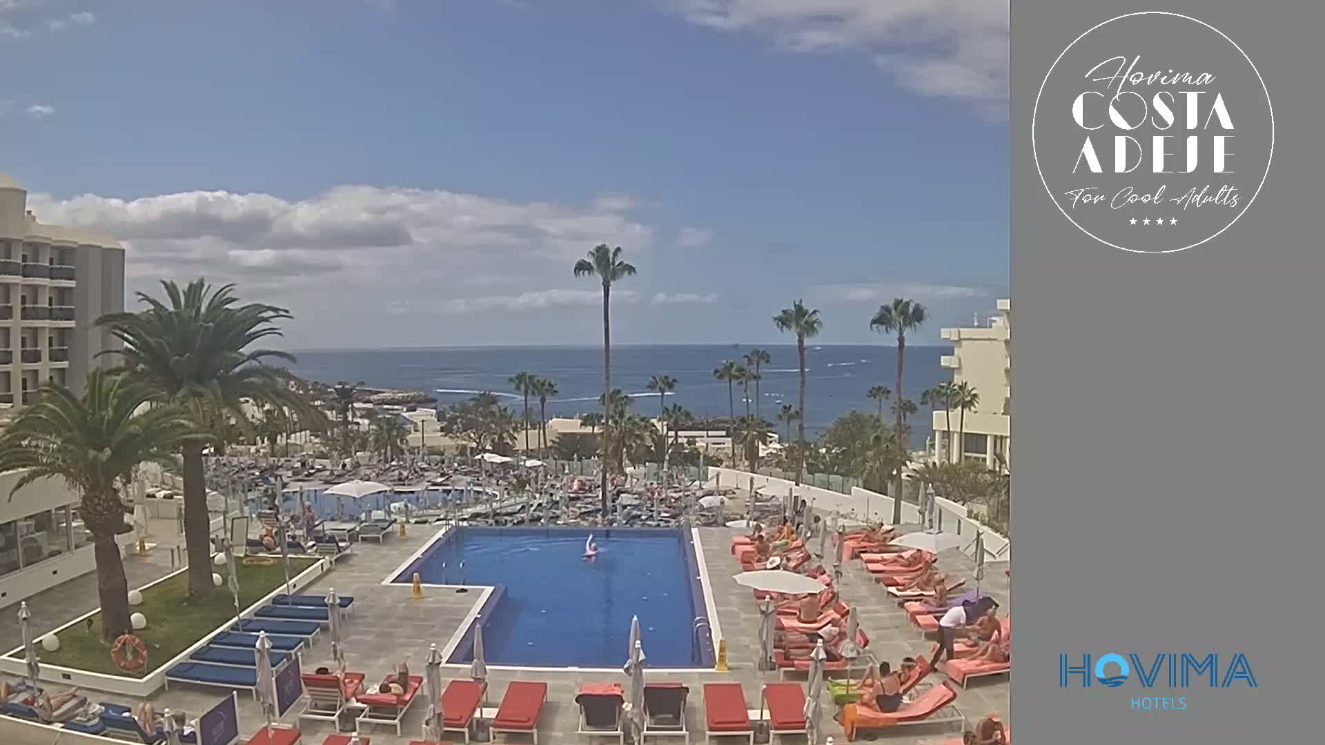 A resort pool area with many lounge chairs and people relaxing under a partly cloudy sky, overlooking a calm ocean.
