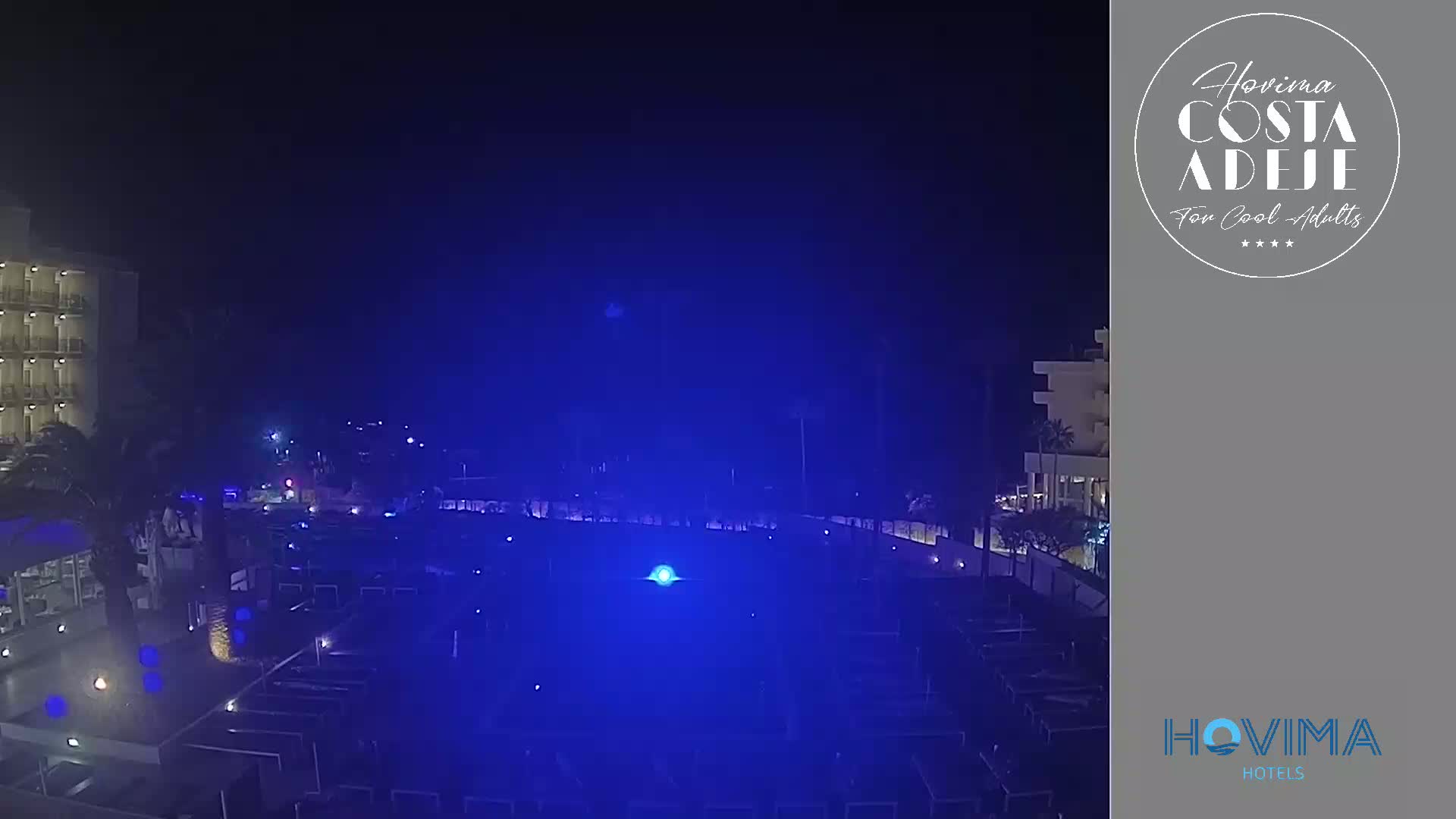 A nighttime view of a resort pool area illuminated in blue light, next to a multi-story building under a dark sky.