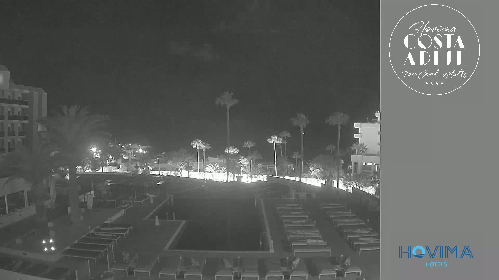 A nighttime, low-light view of a resort pool area with surrounding buildings and palm trees.