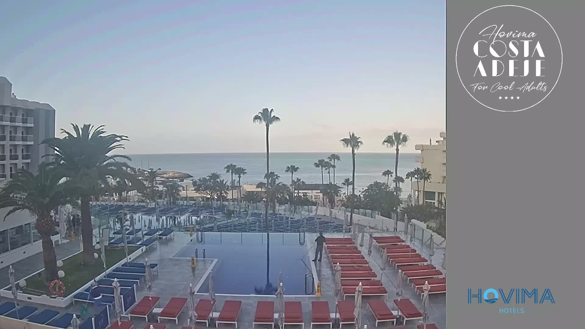 A resort pool area with numerous lounge chairs and umbrellas overlooks a calm ocean under a clear sky.