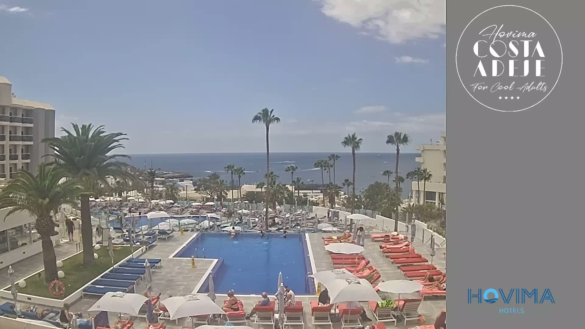 A resort pool area with many lounge chairs and umbrellas surrounding a rectangular pool, overlooks a calm ocean under a mostly sunny sky.