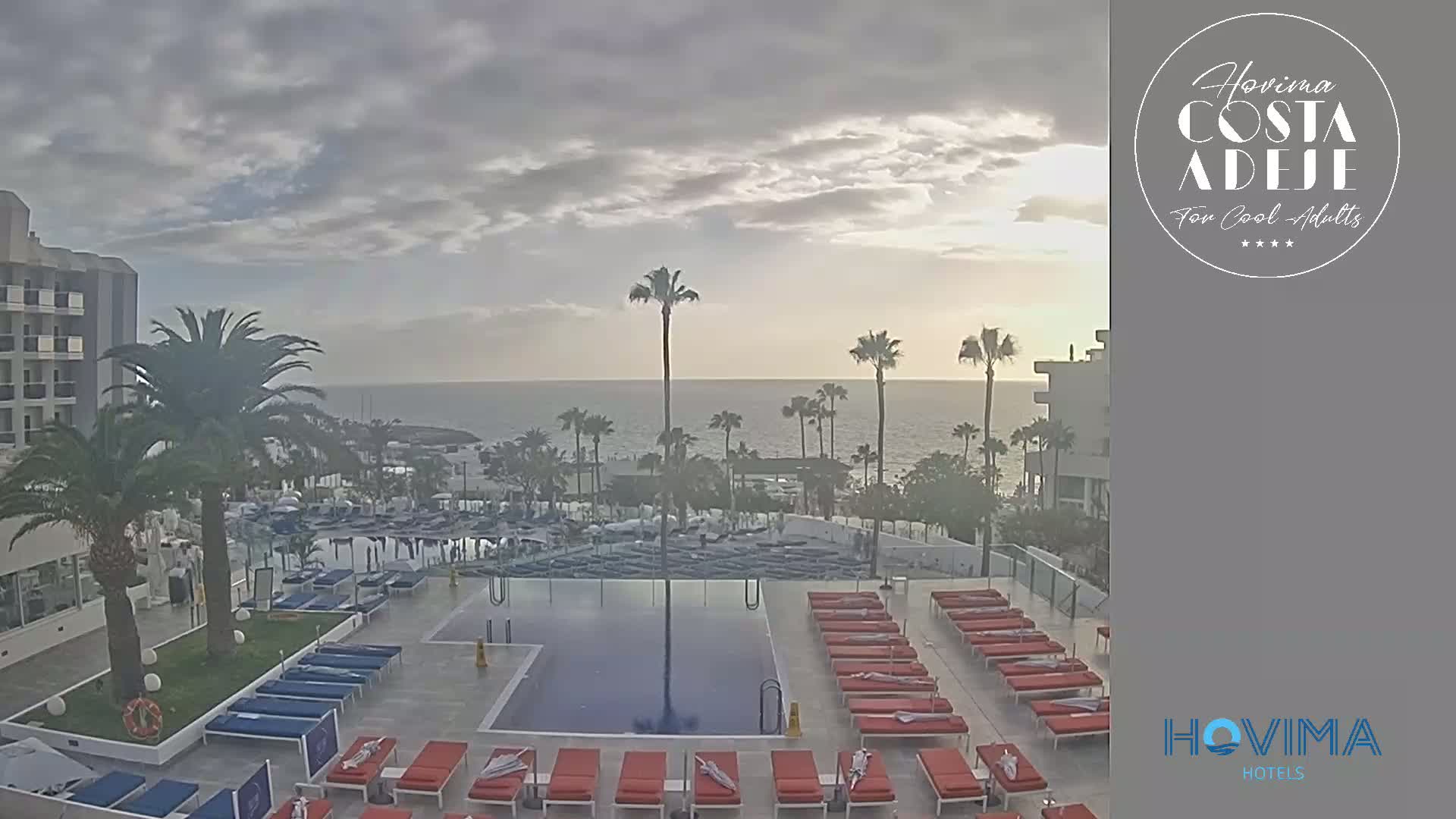 An outdoor pool area with numerous lounge chairs surrounding a rectangular pool, overlooks a calm ocean under a mostly cloudy sky.
