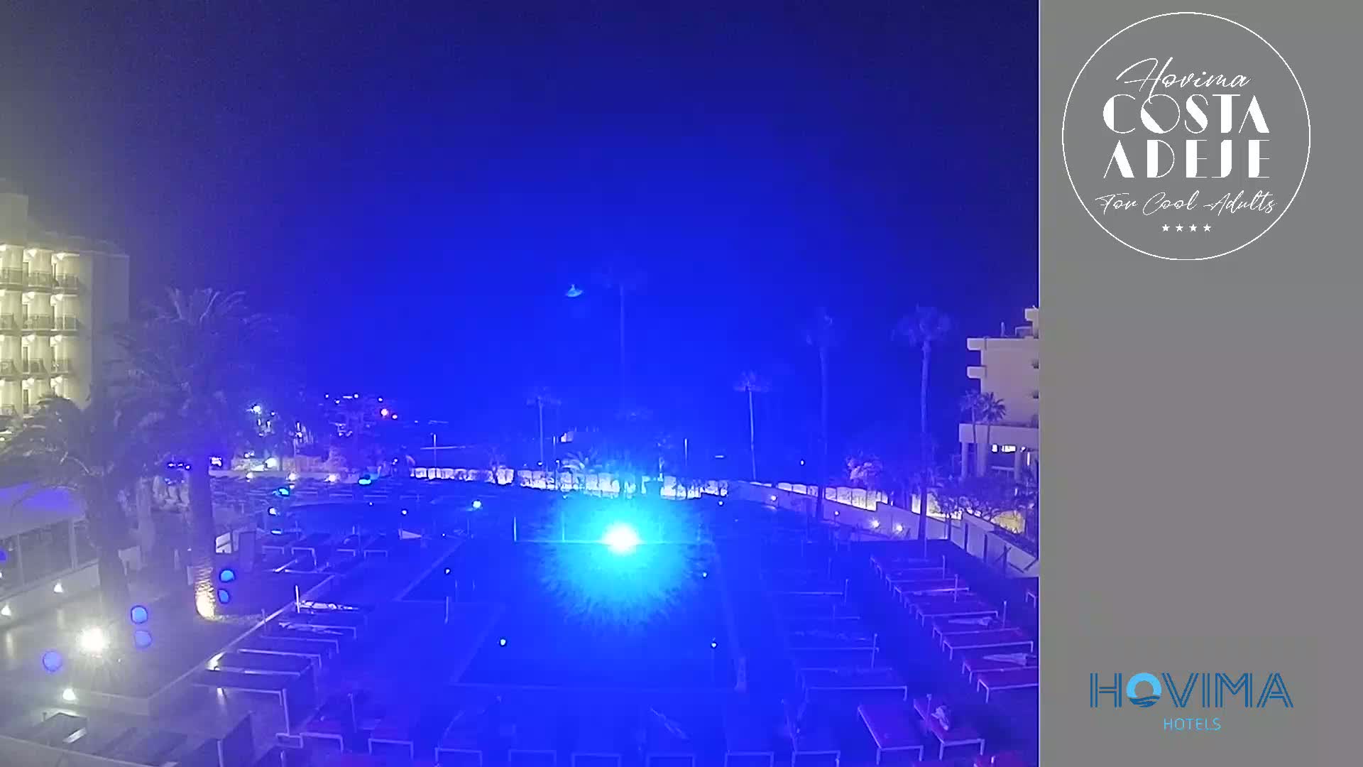 A nighttime view of a resort pool area illuminated in blue light, with palm trees and buildings visible in the background under a dark, possibly hazy sky.