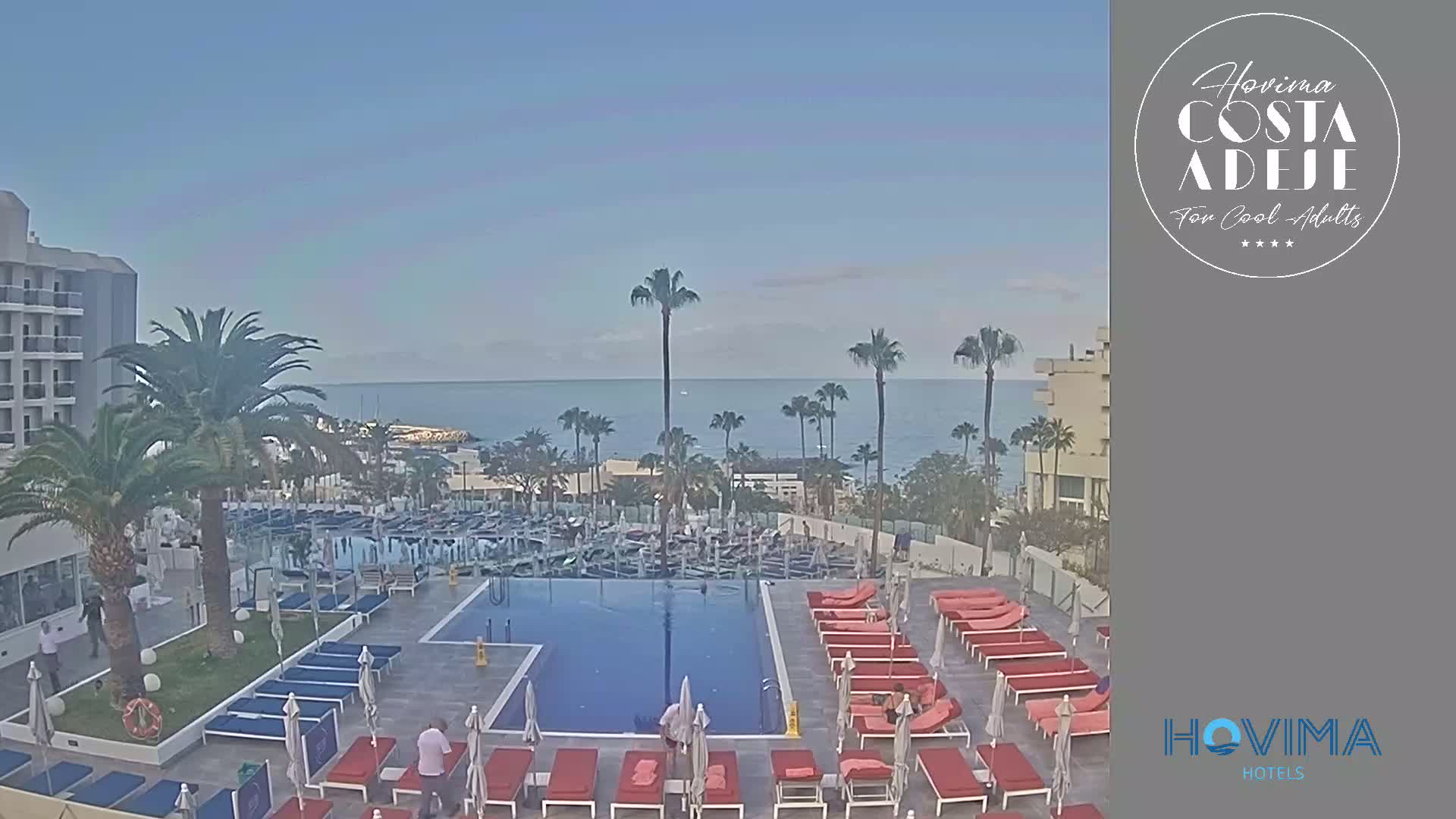 A hotel pool area with red lounge chairs surrounds a blue rectangular pool, overlooking a calm ocean under a clear sky.